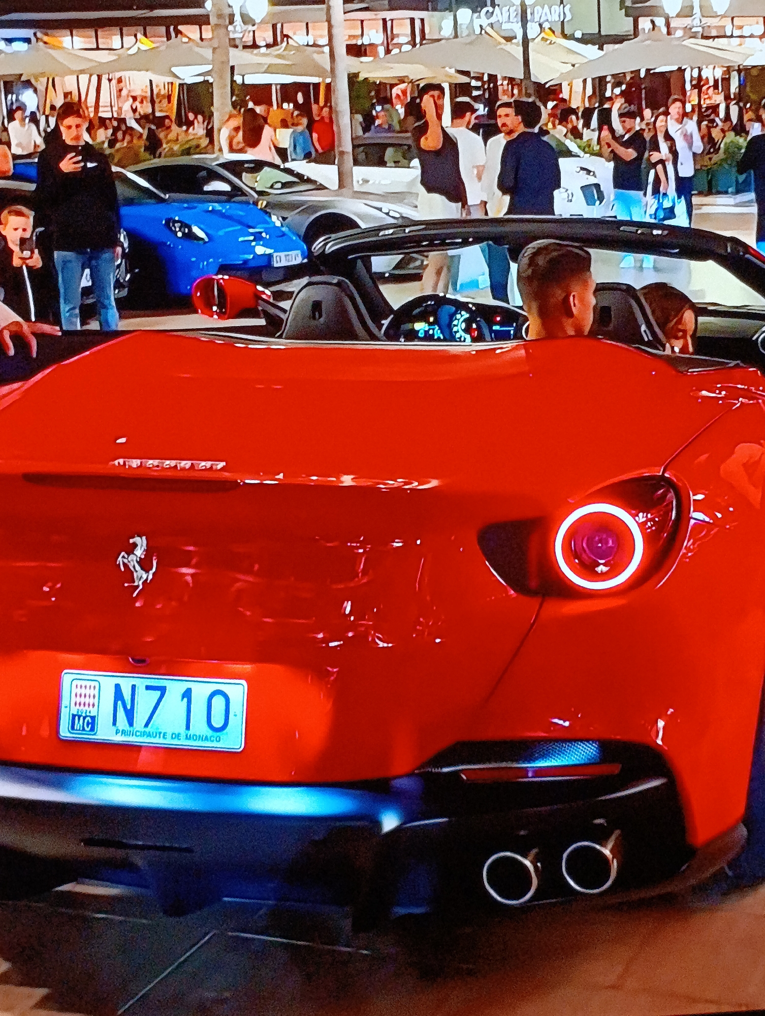 The image shows the rear of a red Ferrari Portofino with Monaco license plate "N710" parked on a street. The background features a crowd of people, with some taking photos of the cars. Visible in the background are other luxury vehicles, including a blue sports car and a light-colored sedan. "CAFE DE PARIS" is visible in the background. The image appears to have been taken in Monte Carlo, Monaco, given the license plate.