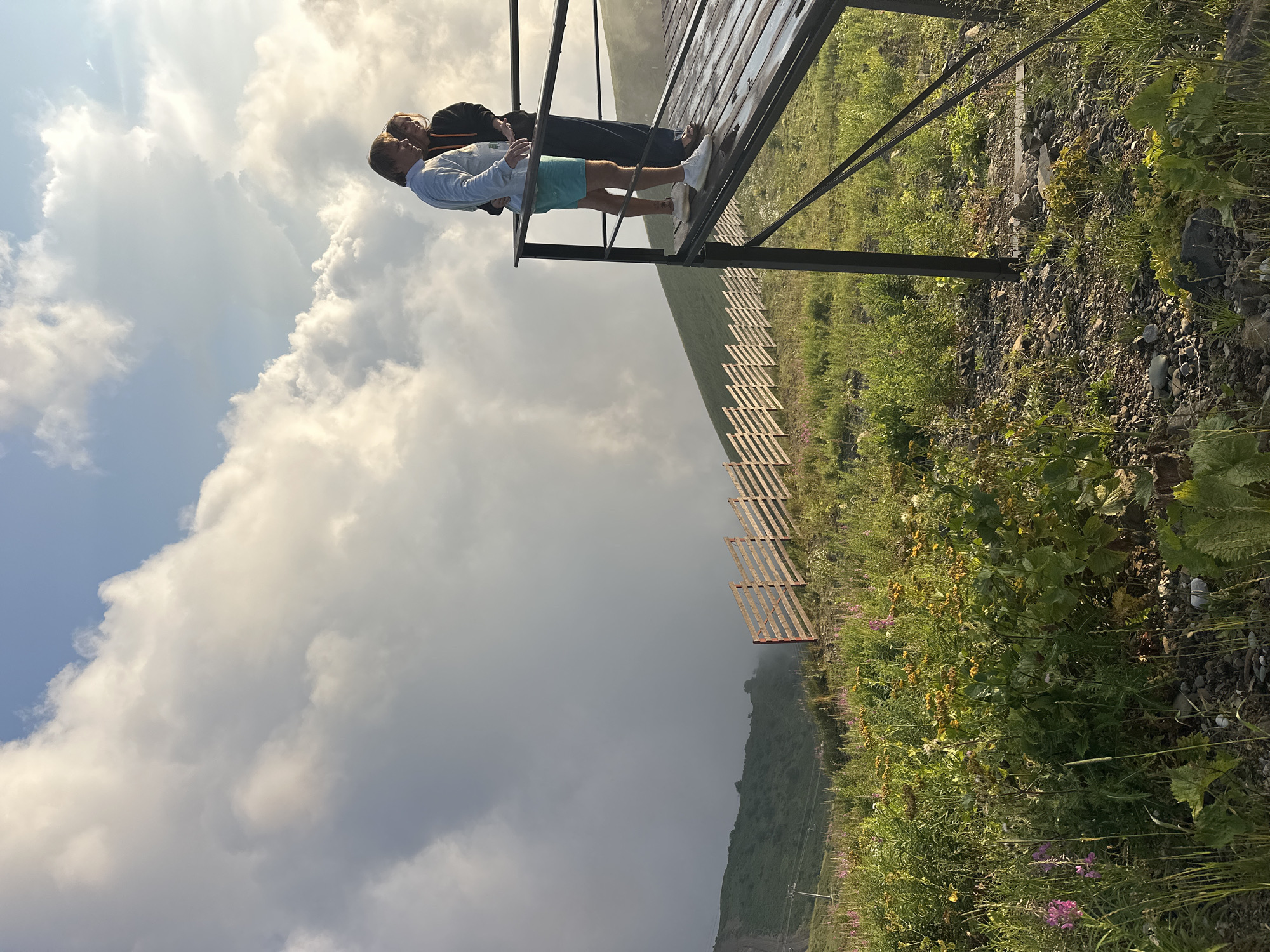 An exterior, medium-shot photograph captures a couple standing on a wooden platform overlooking a mountainous landscape in Estosadok, Russia. The couple, positioned in the upper right quadrant of the frame, is seen from the side as they gaze out at the view. The man, on the left, wears a light blue long-sleeved shirt and turquoise shorts. The woman, on the right, is dressed in a dark hoodie and dark pants. The platform they stand on is weathered, with visible water droplets on its surface, suggesting recent rain.
The background depicts a steep, grassy mountainside receding into the distance. A rustic wooden fence runs diagonally across the midground, marking the edge of a slope. The sky is filled with dramatic, large white clouds, with patches of blue sky visible between them. The foreground is a rocky and grassy terrain dotted with wildflowers, including pink and yellow varieties. The overall scene conveys a serene, natural setting at high altitude.