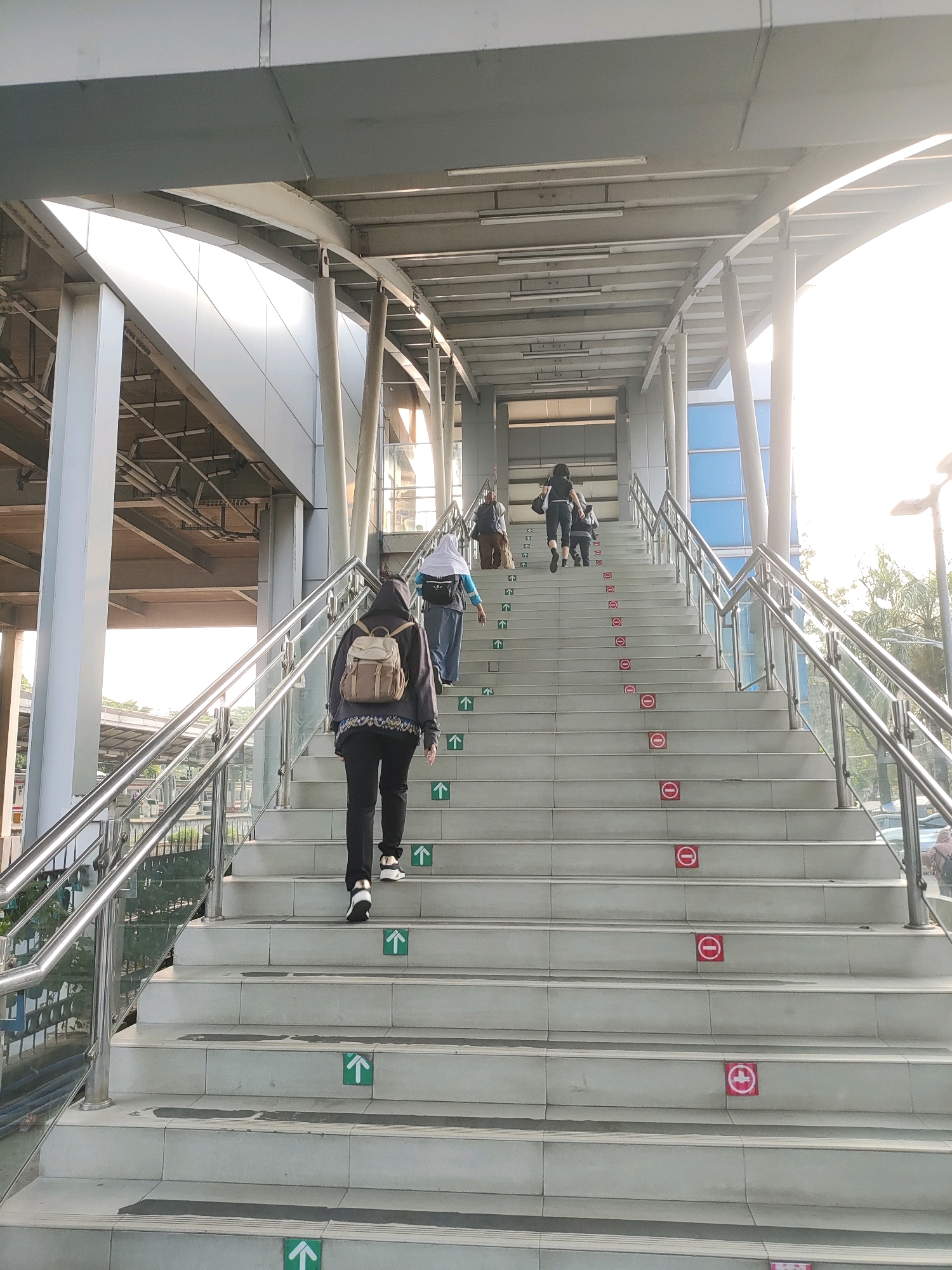 The image shows an outdoor staircase at an elevated transit station in East Jakarta, Indonesia. Several people are ascending the stairs. The person in the foreground, seen from behind, is wearing a dark hooded sweatshirt, black pants, and a light brown backpack. To their left and slightly ahead, a person in a light blue shirt and hijab is also ascending. Further up the stairs, two more individuals are visible. The staircase has silver handrails on both sides and is marked with green directional arrows indicating upward movement on some steps, and red circular signs with diagonal lines, signifying prohibition, on alternating steps. The structure features a modern, metallic roof supported by cylindrical pillars, and sunlight streams in from the right, illuminating the scene.