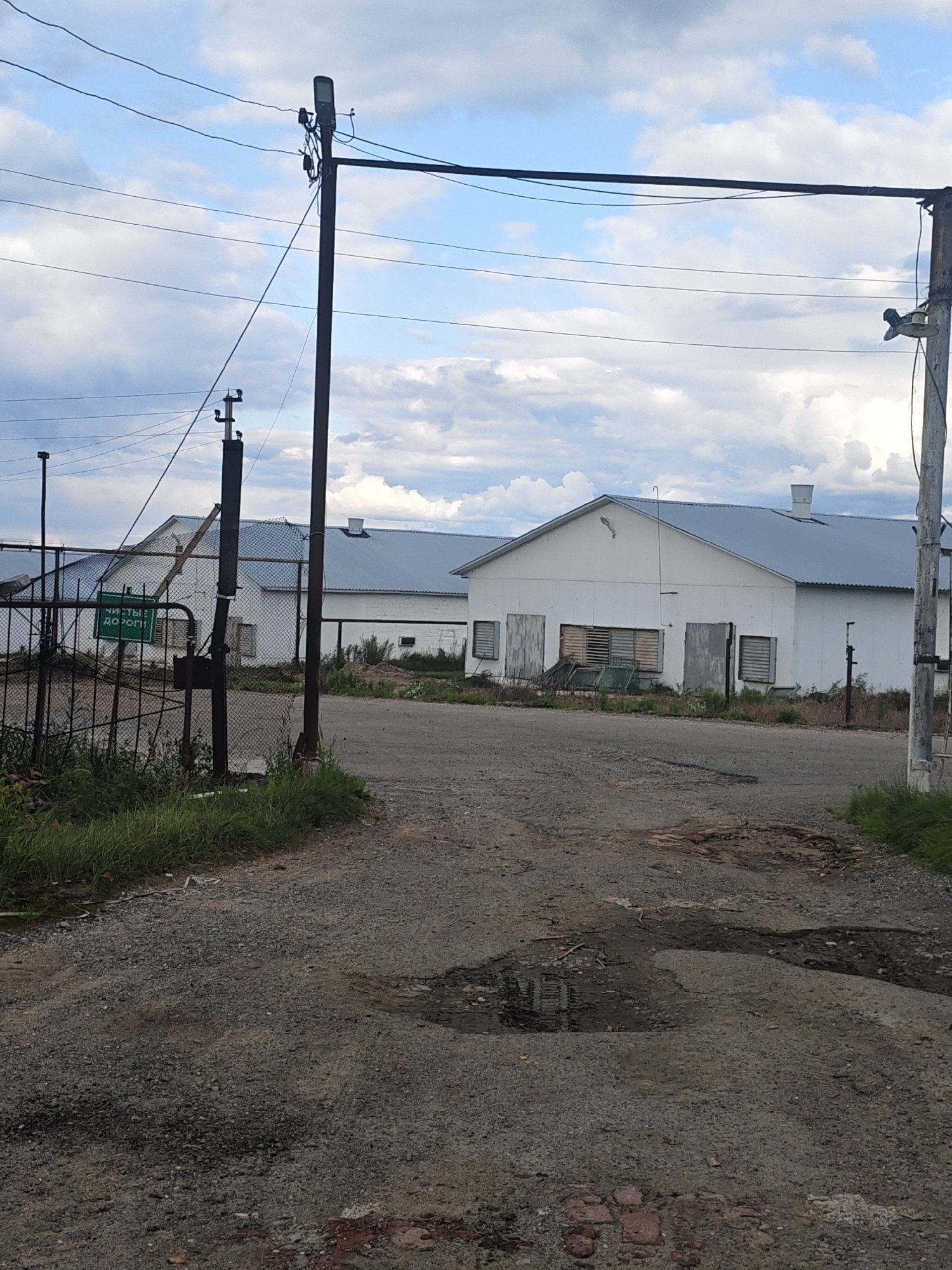 The image shows an outdoor scene in the city of Platoschino, Russia. In the foreground, a gravel road is visible with several puddles and some scattered debris. A metal fence with a green sign that reads "ЧИСТЫЕ ДОРОГИ" (Clean Roads) is on the left side of the road. Beyond the fence are several industrial-looking white buildings with grey roofs. Overhead, power lines and utility poles are present against a cloudy sky. The overall impression is of an industrial or agricultural area.