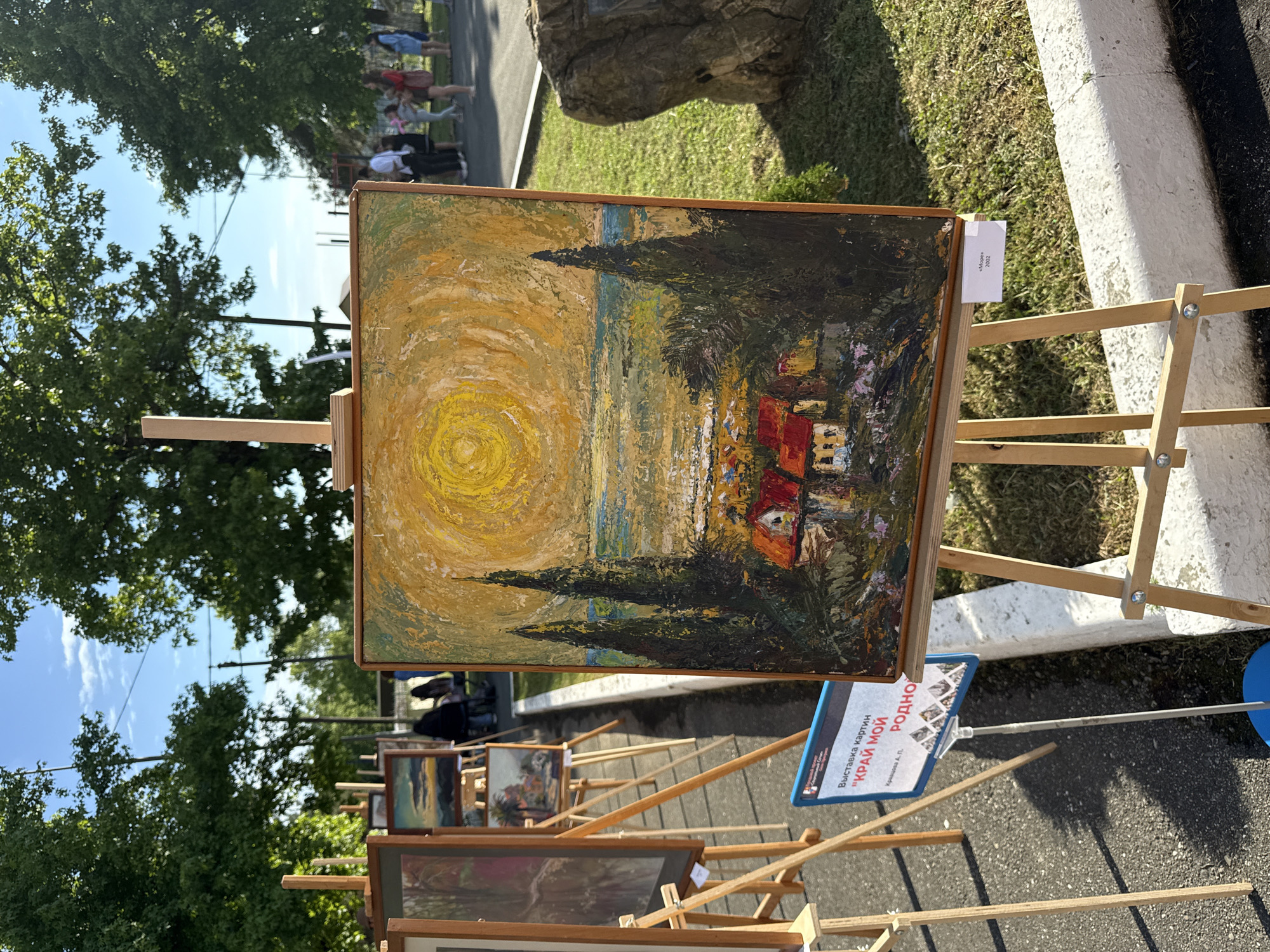 The image depicts an outdoor art exhibition in the city of Progress, Russia. Several paintings are displayed on easels. The central painting, an impressionistic landscape with houses, cypress trees, and a bright yellow sun, is the focal point. In the background, people are seen observing the artworks. The environment includes green trees, a clear sky with some clouds, and a paved walkway. A sign in Russian, identifying the exhibition as "Выставка картин "КРАЙ МОЙ РОДНОЙ"", is visible in the foreground.