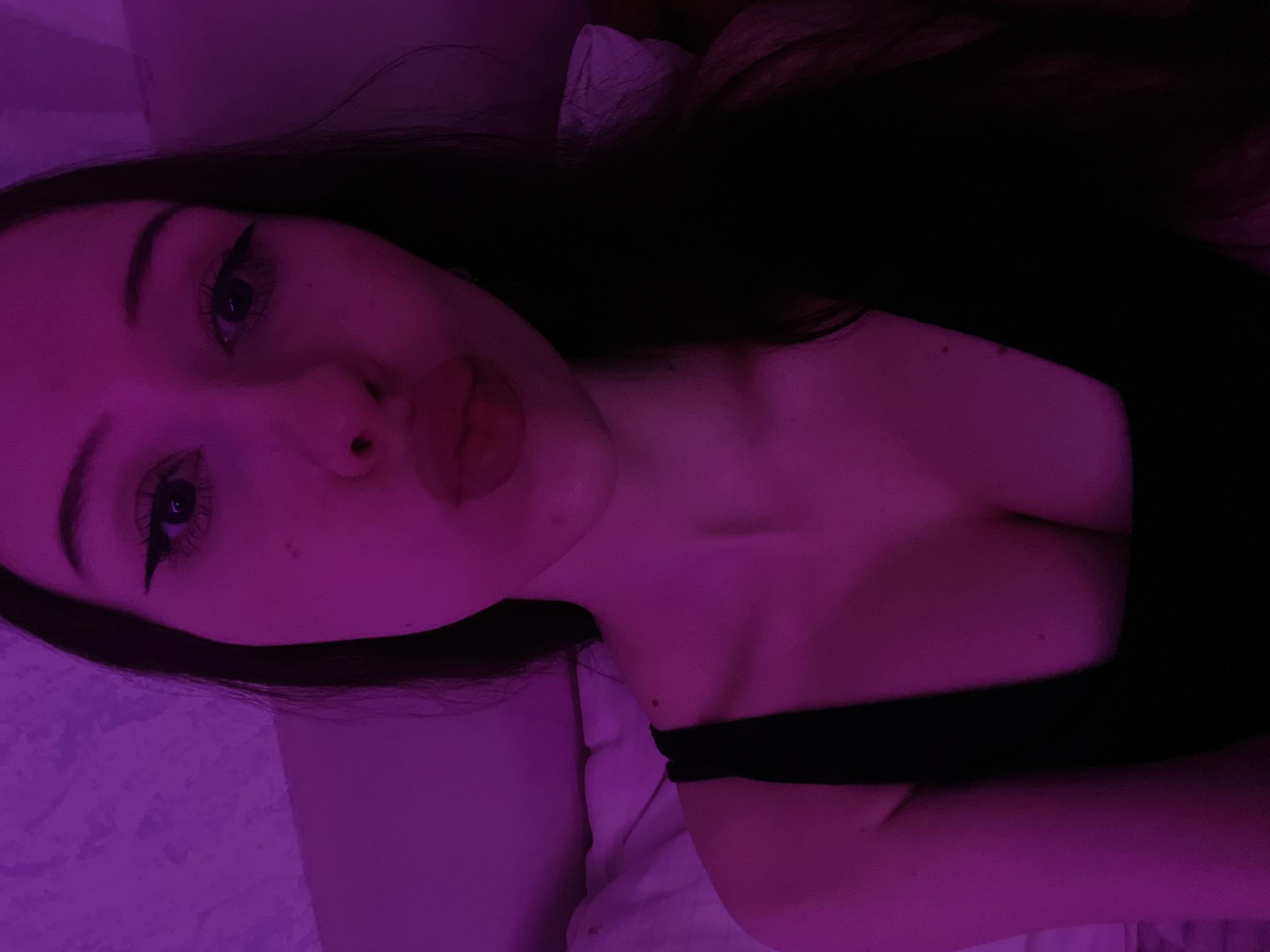 A close-up, indoor, selfie-style photograph features a young woman with long, dark hair. She is wearing a black top with a low neckline, revealing her décolletage. The image is illuminated with purple lighting, casting a pinkish-purple hue over the scene. The woman's face is angled towards the camera, with her lips pursed in a kissing gesture. Her eyes are open, and she has dark eyeliner and mascara. The background appears to be a wall and a bed with white sheets. The image was taken in Votkinsk, Russia.