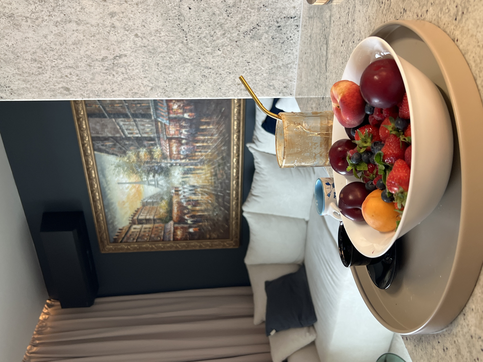 The image captures a cozy interior scene. A fruit bowl filled with strawberries, blueberries, plums, and a peach sits on a tray. A gold straw is visible next to the bowl. In the background, there's a framed painting hanging on a dark wall, and a black speaker stands to the left. A cushioned seating area with multiple pillows is also present. The location is not discernable from the image, so I cannot determine if it is in Plovdiv, Bulgaria.