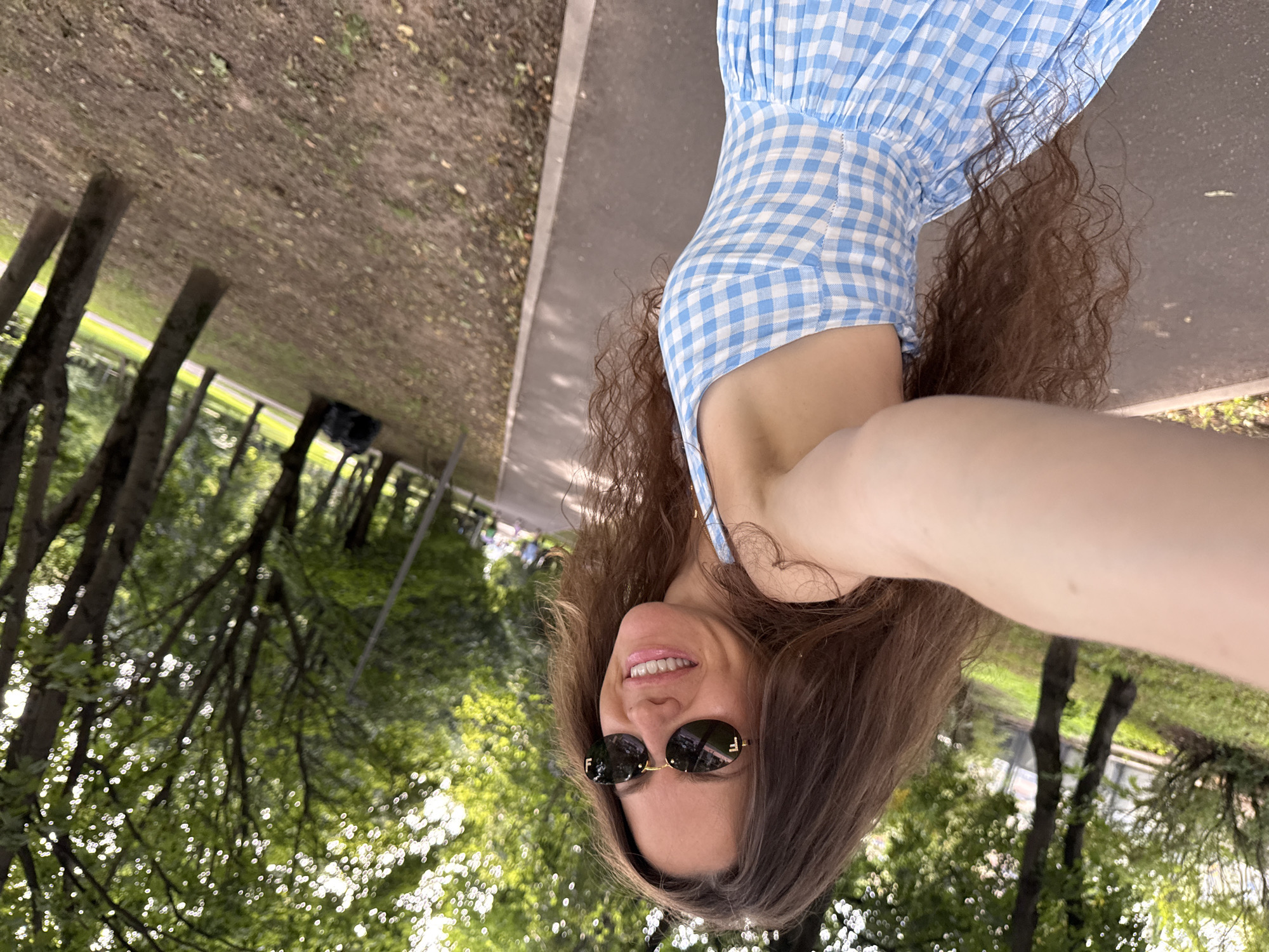 The media file is a selfie taken outdoors in what appears to be a park or a tree-lined walkway. The main subject is a woman with long, wavy brown hair, wearing a blue and white checkered dress and sunglasses. She is smiling at the camera, with her arm extended to take the selfie. In the background, there are trees with lush green foliage and a paved pathway. The sunlight filtering through the leaves creates a dappled effect. The overall mood of the image is cheerful and relaxed. The location is identified as Moscow, Russia. There is no visible text in the media file. The image is rotated 90 degrees counterclockwise.