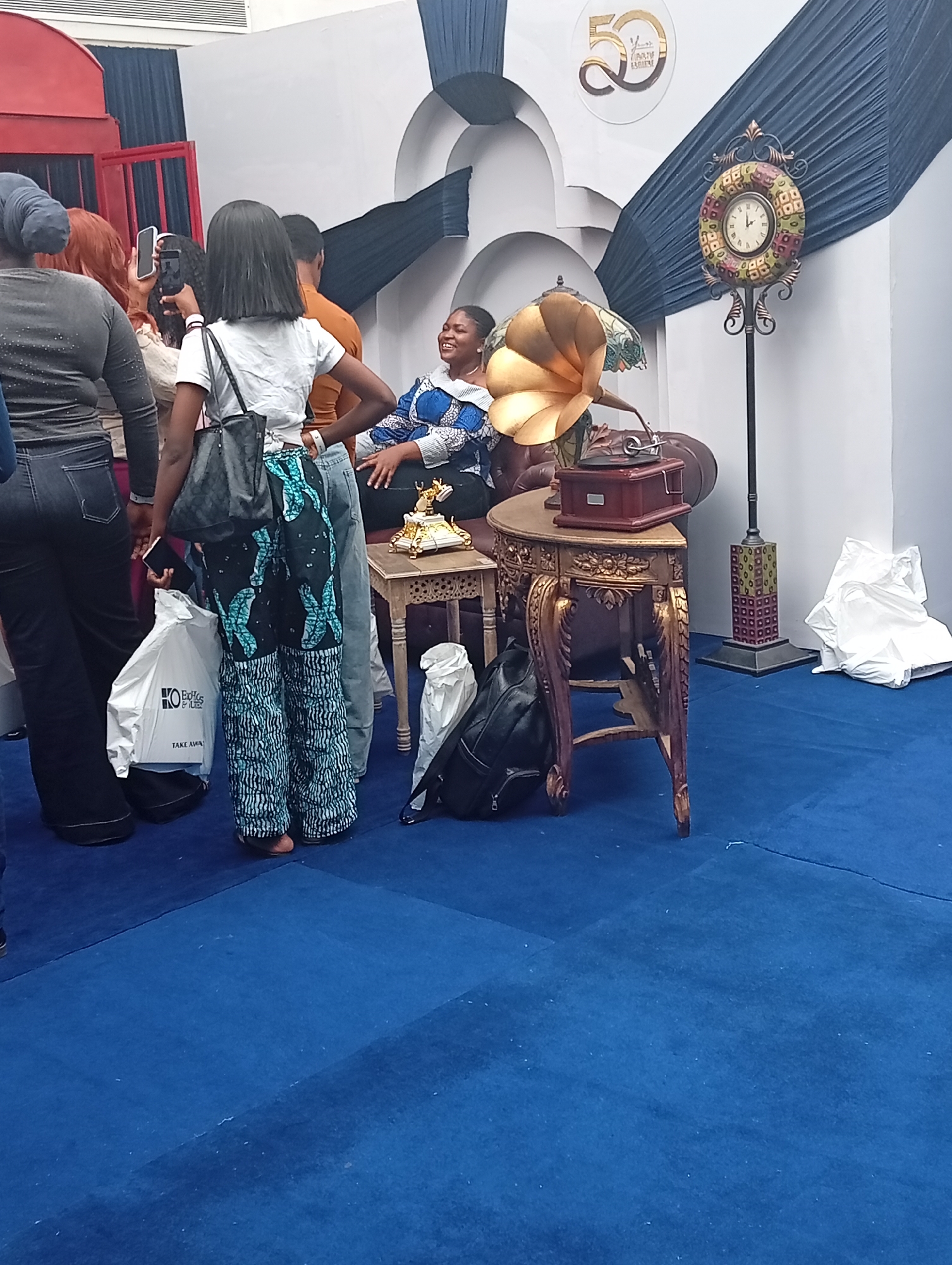 The scene depicts a staged photo opportunity area in Itirin, Nigeria, likely indoors. A woman is seated on a brown leather couch with a golden flower-shaped lamp behind her. A gramophone sits on a small table beside the couch. A decorative clock stands nearby. Several people are gathered around, taking photos with their phones. A white bag with the words "KO Bella by Bell" is visible. The floor is covered with a blue carpet.