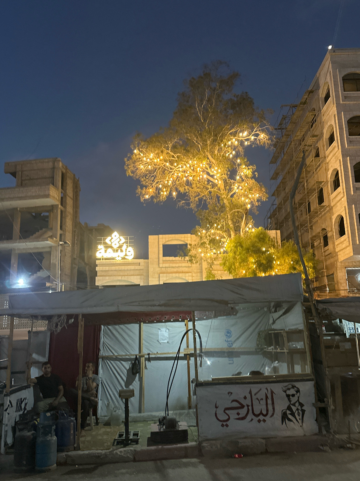 The image depicts an outdoor scene at dusk or night in the city of Gaza, Palestinian Territories. In the foreground, there is a covered stall or makeshift shop with a white tarp roof. Two individuals are seated inside the stall. To the left, there are several gas cylinders. The stall is built against a backdrop of buildings under construction or in a state of disrepair, with exposed concrete and scaffolding visible.
Dominating the midground is a large tree heavily decorated with numerous small, warm-toned lights, giving it a festive or illuminated appearance. Behind the tree, a building with a prominent sign featuring Arabic script and a stylized logo is illuminated. To the right, a multi-story building, possibly an apartment complex or office building, is also visible, with scaffolding suggesting ongoing construction. The sky is a deep blue, indicating late evening. On the front of the stall, there is text in Arabic script and a black and white drawing of a person with distinctive hair and sunglasses.