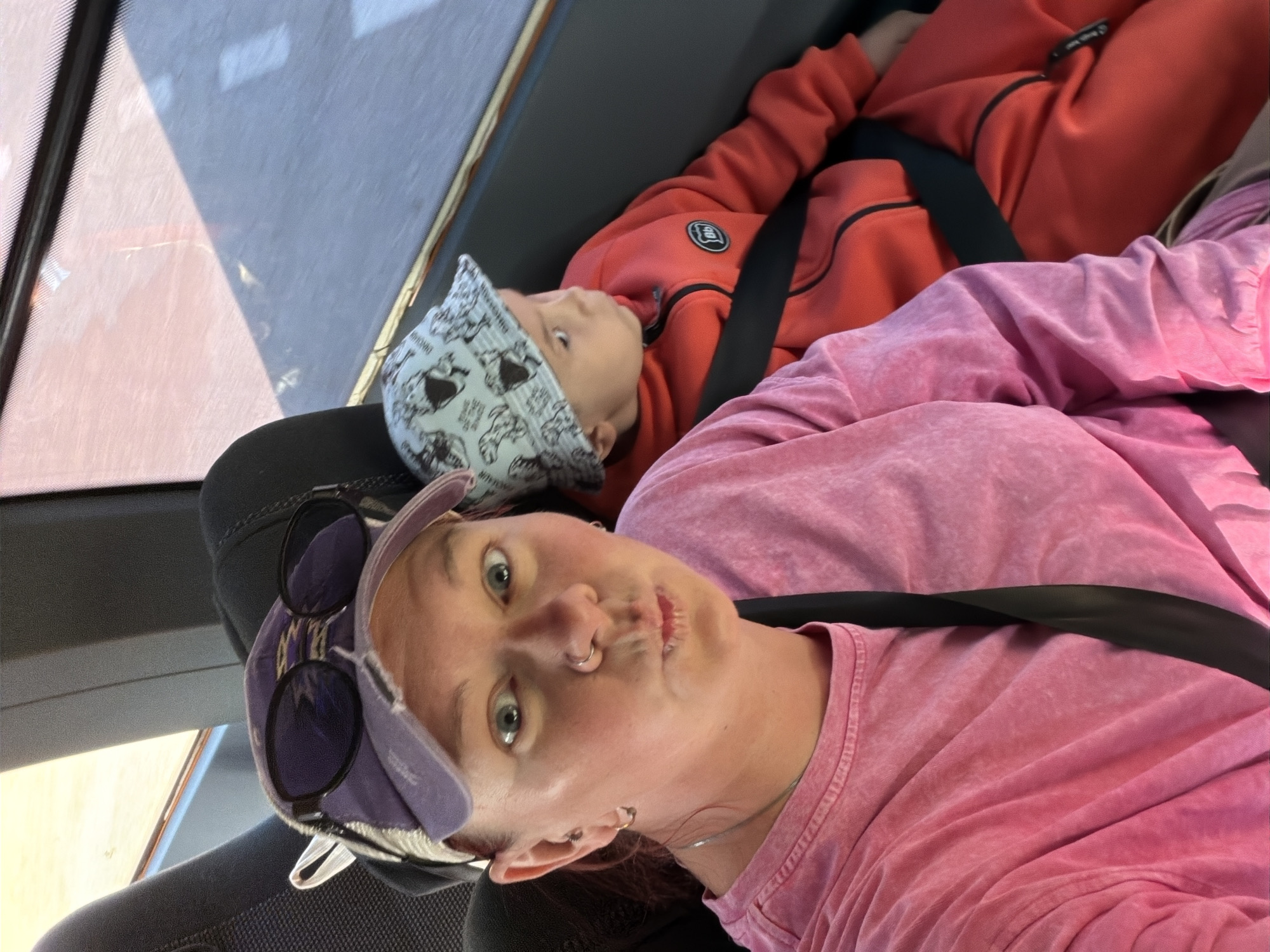 The image shows a selfie taken inside a car. A woman with fair skin, wearing a pink shirt and a purple baseball cap with sunglasses on top, is looking at the camera. She has a nose ring and earrings. A young child wearing a red jumpsuit and a patterned hat is visible behind her, lying down and strapped into the seat with a seatbelt. The car's interior is visible, including parts of the window and the seat. The location is not discernible from the image. The image does not explicitly indicate the city of Chaikovsky, Russia.