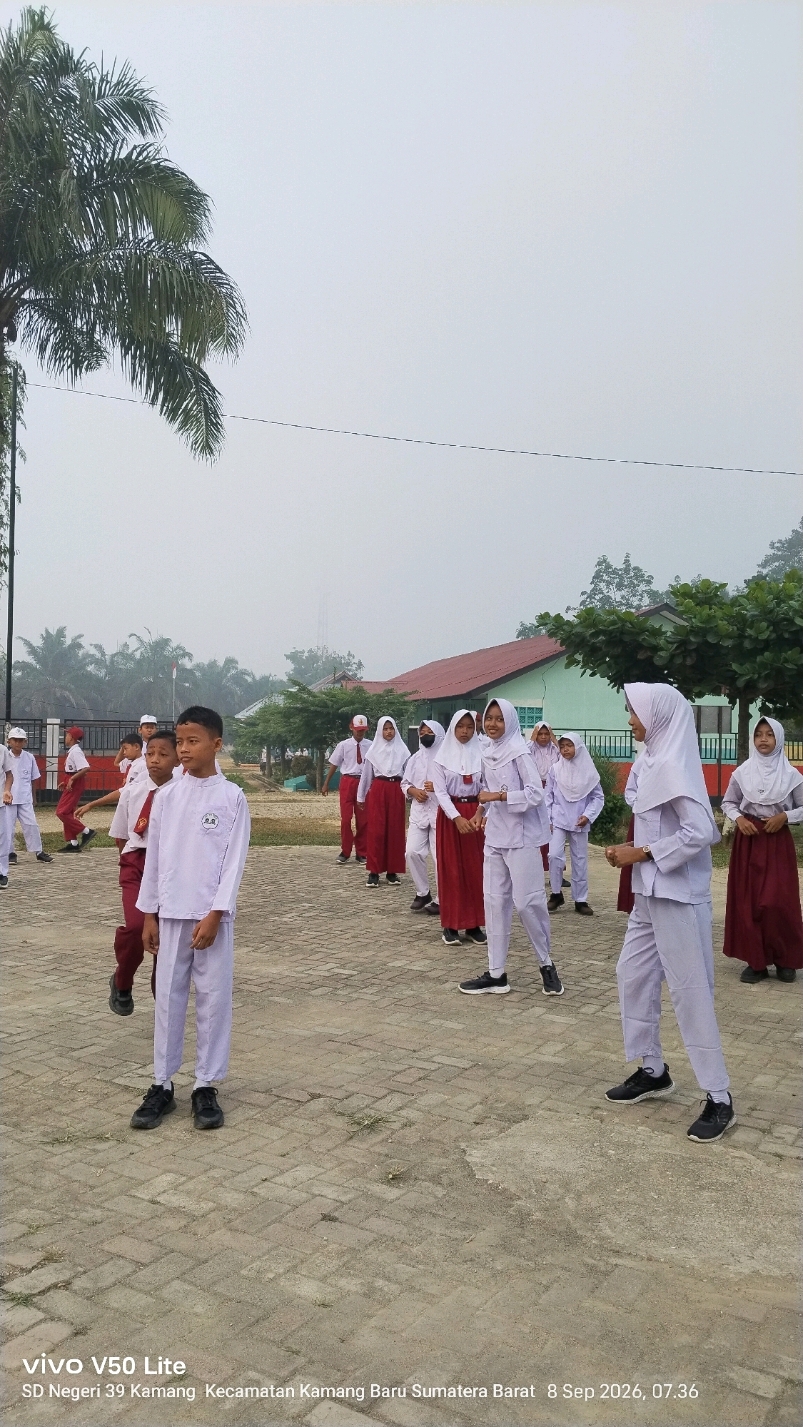 The image shows a group of students in uniform standing outdoors in what appears to be a schoolyard. The students are of various ages and are wearing white shirts and pants or white shirts and maroon skirts. Some of the students are wearing headscarves. The ground is paved with interlocking bricks. In the background, there are buildings with red roofs and green walls, trees, and palm trees. The sky is overcast. A watermark at the bottom of the image indicates the location as "SD Negeri 39 Kamang Kecamatan Kamang Baru Sumatera Barat" and the date and time as "8 Sep 2026, 07.36".