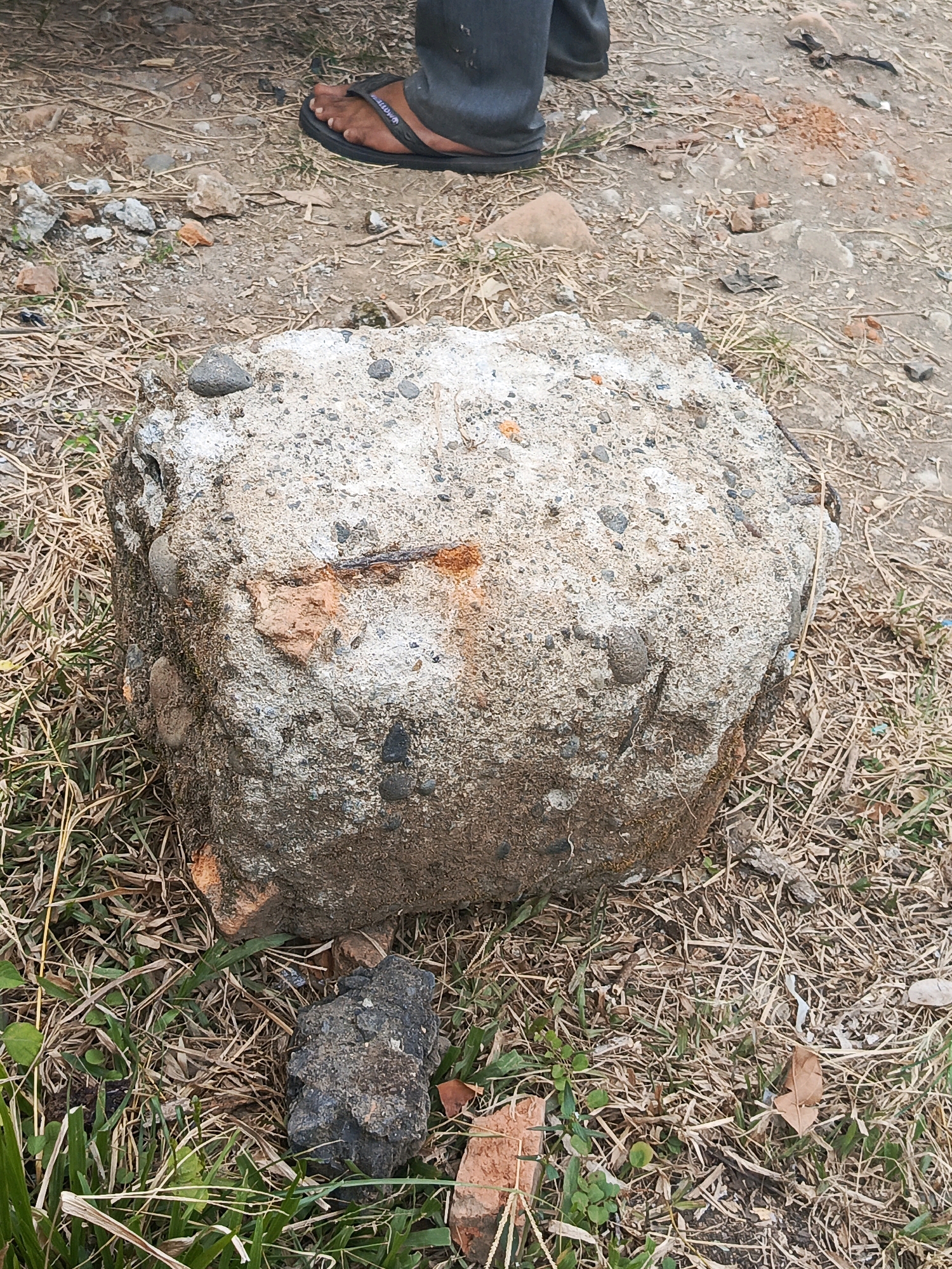A close-up, eye-level shot features a large, irregularly shaped concrete object resting on dry grass and dirt. The concrete is a mix of grey cement, small pebbles, and larger stones, with visible patches of rust-colored material, possibly brick fragments or oxidized metal. A bent piece of rebar protrudes from the top surface. The ground around the concrete is scattered with small stones, dry grass, and some green shoots of grass. In the background, the lower legs and feet of a person wearing dark pants and black flip-flops are visible. The overall environment appears to be an outdoor, possibly construction or debris-filled area in Megang Sakti II, Indonesia.