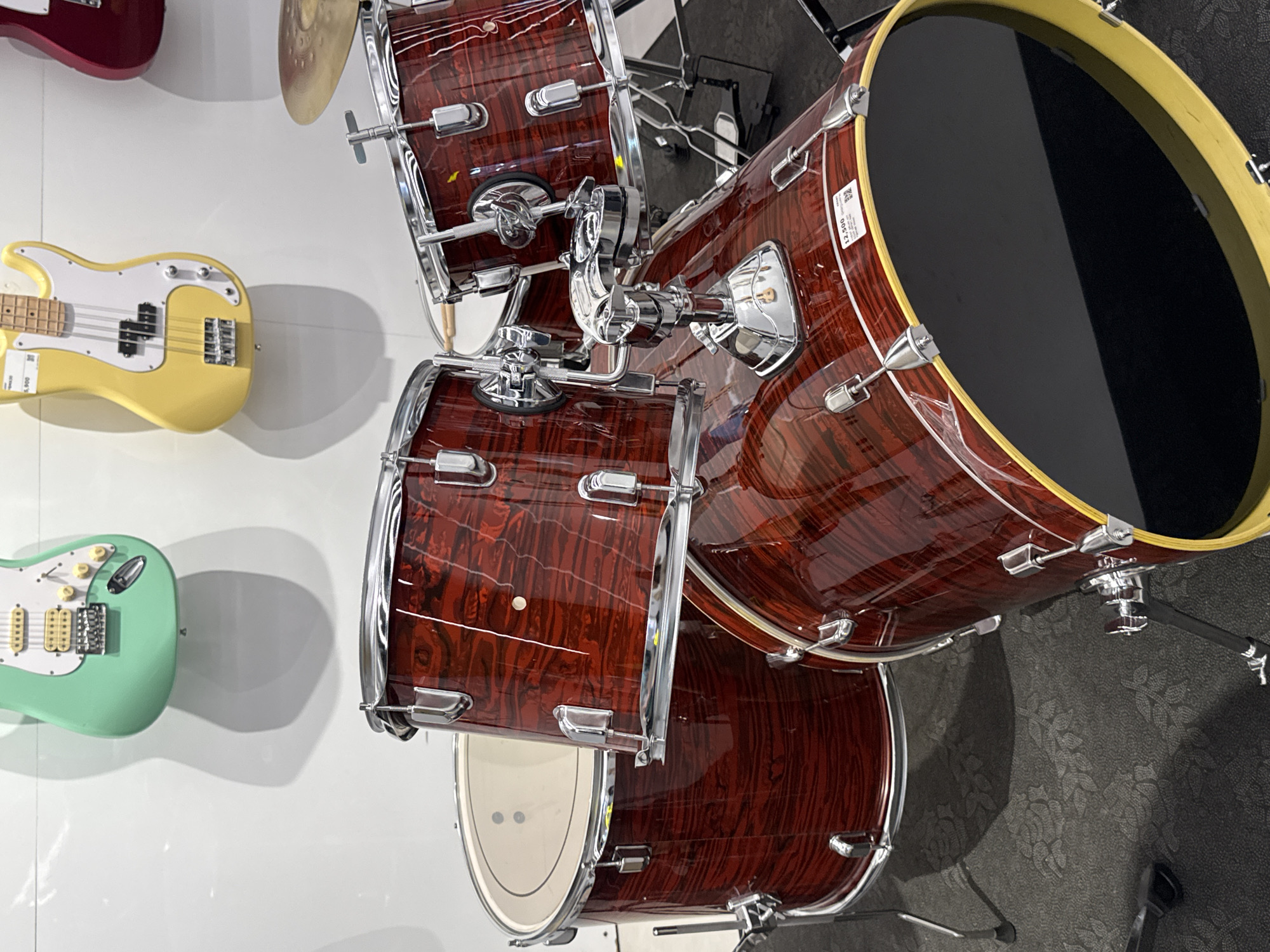 The image shows a retail display of musical instruments, primarily a drum kit and electric guitars, likely within a music store or showroom. The overall scene suggests an indoor environment with clean lighting.
On the right side of the frame, a glossy dark red drum kit with a distinct wood grain finish and gleaming chrome hardware is prominently featured. The large bass drum, positioned towards the upper right, has a black drumhead framed by a yellow hoop. A white sticker with the number "12,500" is visible on the bass drum. Several tom-toms, also in the matching dark red finish, are part of the kit, with their chrome mounts and hoops clearly visible. A portion of a brass cymbal can be seen in the top right corner. The drum kit rests on a dark gray carpet with a subtle floral pattern.
On the left side, against a clean white wall, three electric guitars are displayed. A bright yellow electric bass guitar, styled like a classic precision bass with a white pickguard and a maple neck, hangs centrally. A white price sticker on its body shows "5,500". Below it, a mint green electric guitar, resembling a Fender Stratocaster, with a white pickguard and cream-colored pickups, is also on display. In the upper left corner, part of a dark red electric guitar is visible, mostly cropped out of the frame.
No people are present, and no activity is taking place; the instruments are arranged for viewing or sale. The visible price tags ("5,500" and "12,500"), given the provided location context of Davao City, Philippines, likely represent prices in Philippine Pesos.