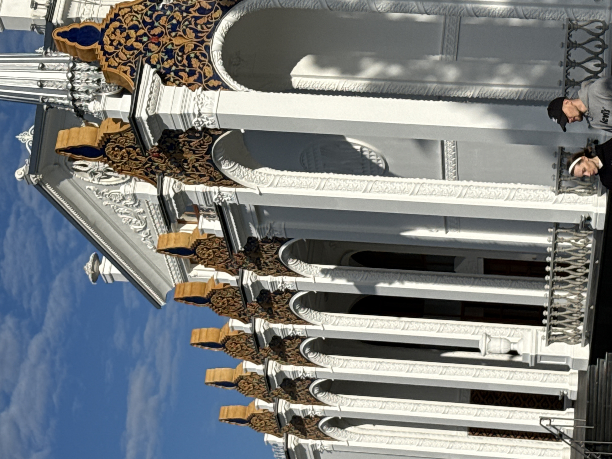 The image captures a bright, sunny day with a clear blue sky filled with wispy clouds. The scene focuses on the ornate exterior of a building, likely a pavilion or exhibition hall, characterized by white columns, arched entrances, and elaborate decorative elements. These decorations feature rich blue and gold patterns with intricate floral motifs, topped with golden finials.
In the foreground, two individuals are visible. A woman on the left wears a black hoodie and a white headband. A man on the right wears a grey hoodie with "Levi's" branding and a black baseball cap. They are walking or standing near the building, seemingly interacting with each other. The architecture suggests a classical or neoclassical style, possibly with Eastern influences due to the decorative patterns. The building's façade is highly detailed, with textured surfaces and sculptural elements. Shadows cast by the sun highlight the architectural features and the presence of the people.
This building is located in Moscow, Russia, and is identified as the Armenian Pavilion at VDNKh (Exhibition of Achievements of National Economy). The VDNKh is a vast exhibition center in Moscow, featuring numerous pavilions showcasing the achievements of various Soviet republics and industries. The Armenian Pavilion, specifically, is known for its distinctive architecture that blends classical styles with Armenian motifs.