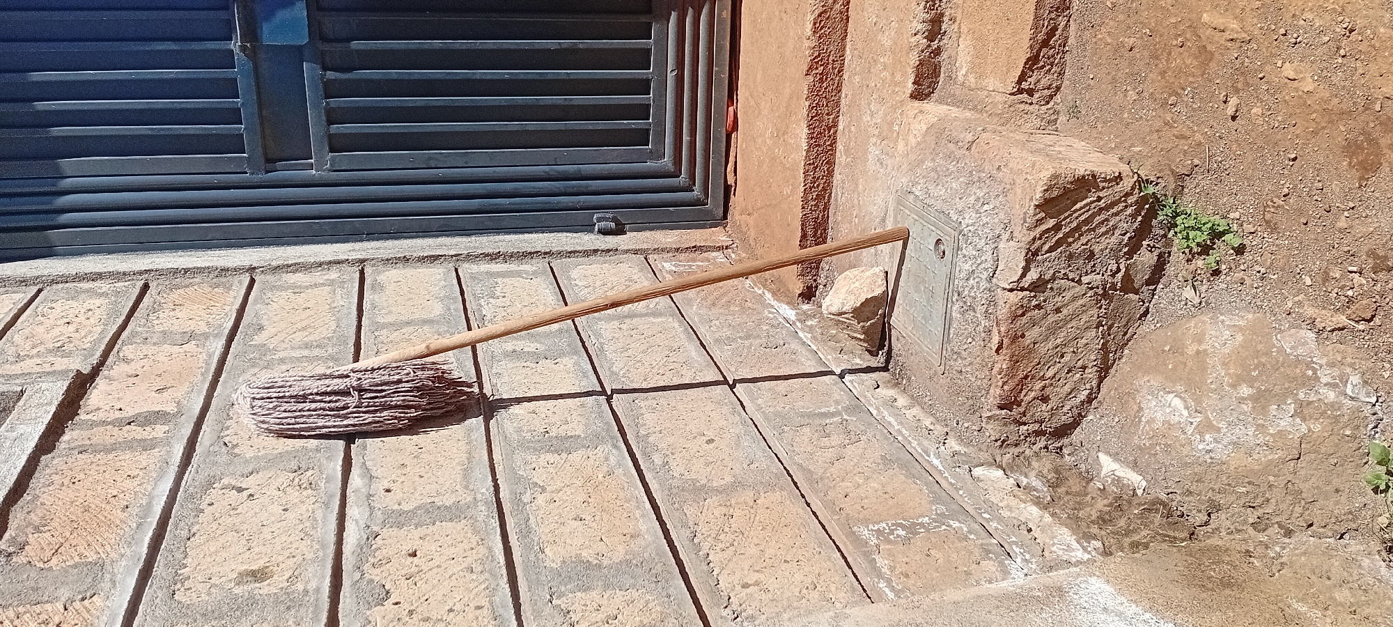 A mop with a wooden handle leans against a textured, light-colored brick wall. The mop head appears to be made of frayed white and pinkish strands. To the right of the wall is a dark blue metal gate with vertical slats. In front of the wall and extending towards the gate are steps made of rough, earthy-colored material. A small, light-colored rock rests on the second step. A metal cover with a grid pattern is visible on the first step. The ground around the steps is composed of dirt and small stones, with a few green plants growing in patches. The environment suggests an outdoor setting, possibly a residential or commercial property.