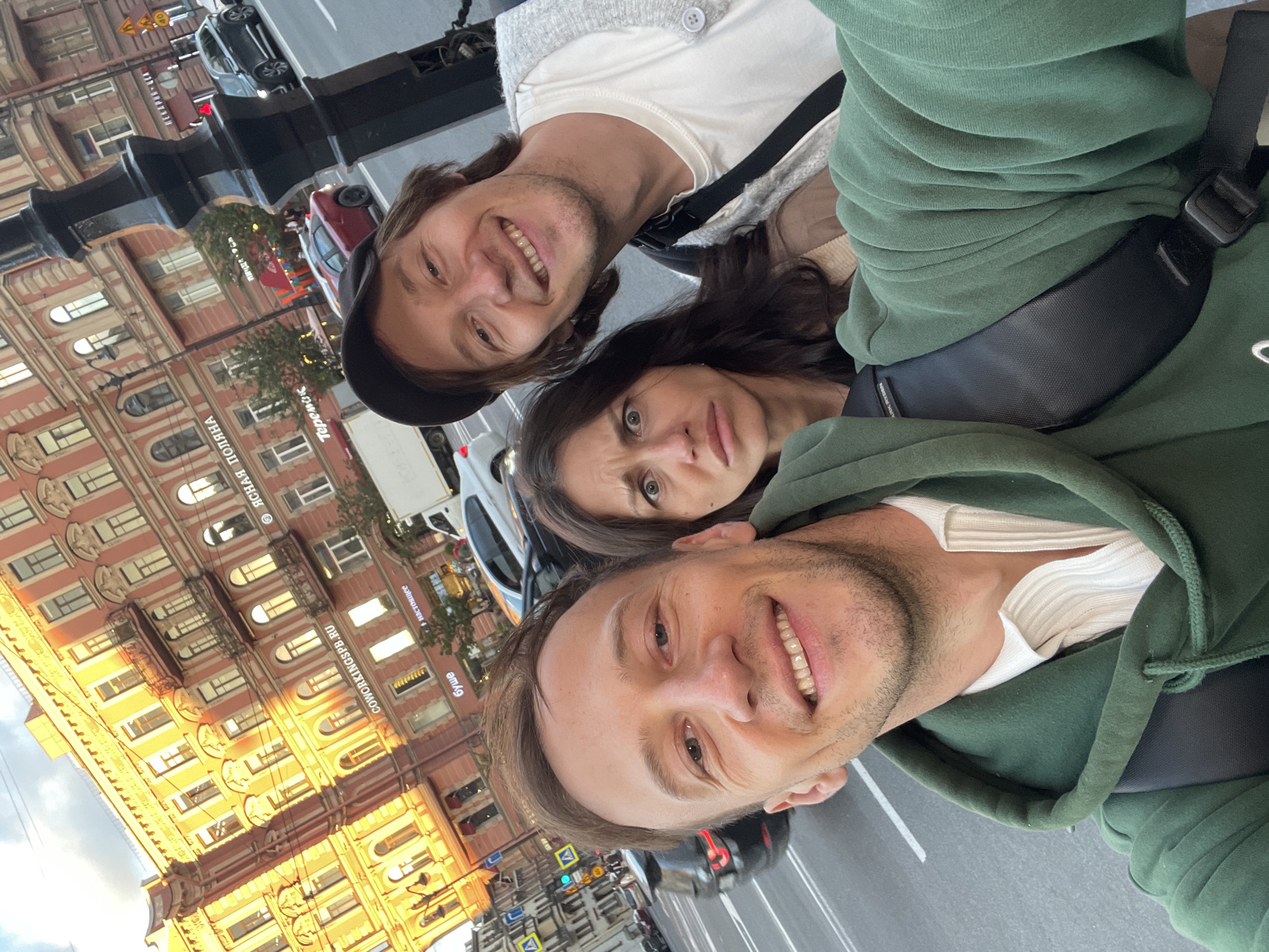 In a selfie taken on a street in Saint Petersburg, Russia, three people are depicted. The person in the foreground, on the left, is a man with fair skin, short brown hair, and a beard, smiling at the camera. He is wearing a green hooded sweatshirt over a white collared shirt. To his right and slightly behind him is a woman with dark brown hair, making a pained facial expression. She is wearing a beige jacket over a tan sweater. Behind her and to the right is another man, also with fair skin and brown hair, wearing a black baseball cap, a white t-shirt, and a grey cardigan. He is smiling broadly at the camera.
The background features a multi-story building with ornate architecture, characteristic of Saint Petersburg, illuminated by the warm light of what appears to be late afternoon or early evening sun. Signs on the building are visible, including "COWORKINGS PB.RU" and "Ясная Поляна". The street in front of the building is paved and has traffic lanes marked. Several cars are visible on the street, including a black SUV and a red sedan. A black lamppost is prominently positioned to the right of the frame. The overall impression is of a candid moment captured during an outing in the city.