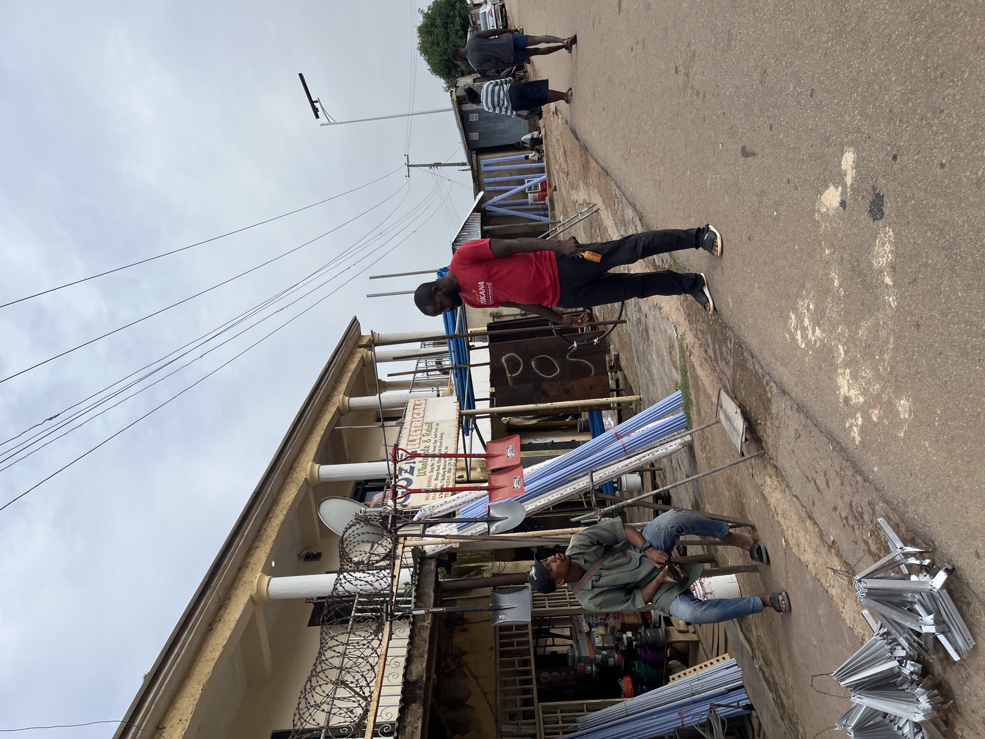 The image, captured in Gura Top, Nigeria, depicts a street scene on an overcast day. The orientation of the image is rotated 90 degrees counter-clockwise. In the foreground, a man in a red t-shirt with "TIKANA" printed on it stands next to a pile of metal shelving brackets. He is holding a small object in his right hand. To his left, a building with white pillars and barbed wire fencing is visible. Further down the street, another man in a green shirt and blue jeans is seated, looking upwards and holding a mobile phone. Various construction materials, including blue pipes and red shovels, are stacked along the sidewalk. In the background, other people are walking along the paved road, and a few buildings line the street. The overall atmosphere suggests an active marketplace or commercial area. Visible text includes "ELECTRICALS", "Wholesale & Retail", "TIKANA", and "POS".