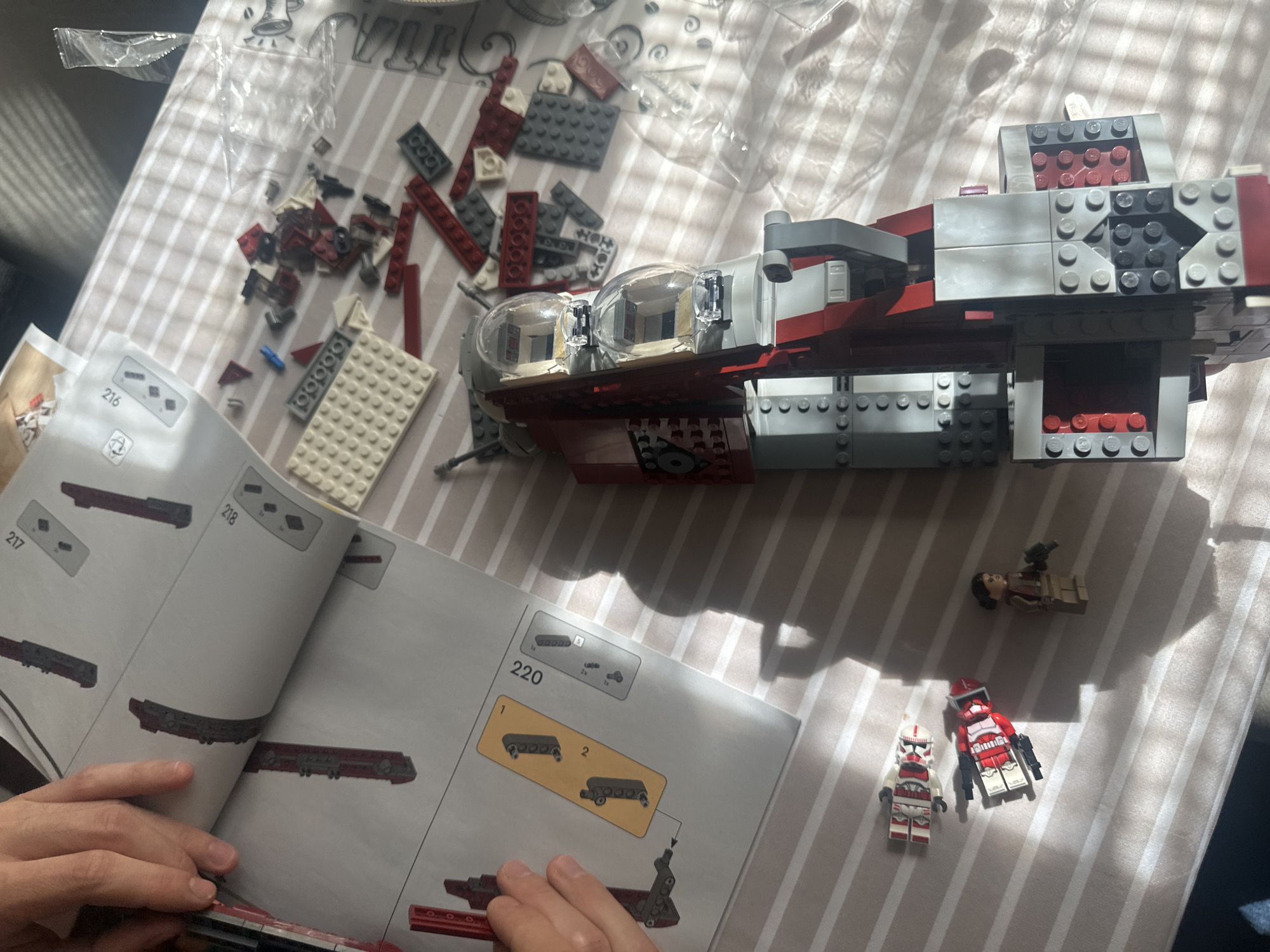 The image captures an indoor scene in Sofia, Bulgaria, focusing on the activity of building a Lego set. A pair of hands is visible at the bottom, holding an open instruction manual and interacting with the Lego pieces.
The main subject is a partially assembled Lego model, which strongly resembles a Republic Gunship from Star Wars, characterized by its grey and dark red color scheme and distinctive dual bubble cockpits. Loose Lego bricks in red, dark grey, white, black, and a few blue pieces are scattered on a striped fabric surface to the left, along with empty clear plastic bags.
The instruction manual is open to pages 216, 217, 218, and 220, displaying various steps and components for the build. Page 220, in particular, shows step 1 and 2 for assembling a part, with piece counts like "1x", "2x", and "5" visible.
To the right of the main model, three Lego minifigures are placed on the striped surface. Two are Clone Troopers: one predominantly red with white accents, and another with a white torso and red markings, both holding black blasters. The third minifigure has a brown torso, dark hair, and a weapon attached to its back.
The setting appears to be a table or similar flat surface covered with a white and grey striped tablecloth or bedsheet. Strong natural light streams in from the top left, casting distinct shadows across the scene, suggesting it is daytime, possibly late morning or afternoon. The overall impression is one of focused construction and a hobbyist's workspace.