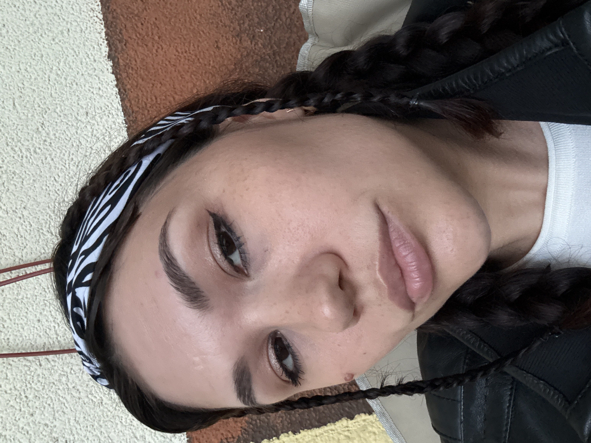 An outdoor, close-up, eye-level, medium-shot photograph shows the face of a young woman with dark hair in braids and a zebra-print bandana. She is wearing a white t-shirt and a black leather jacket. Her eyes are dark brown and she has black eyeliner. She is looking into the camera with her mouth slightly open. The background consists of a textured wall with sections of off-white and brown.