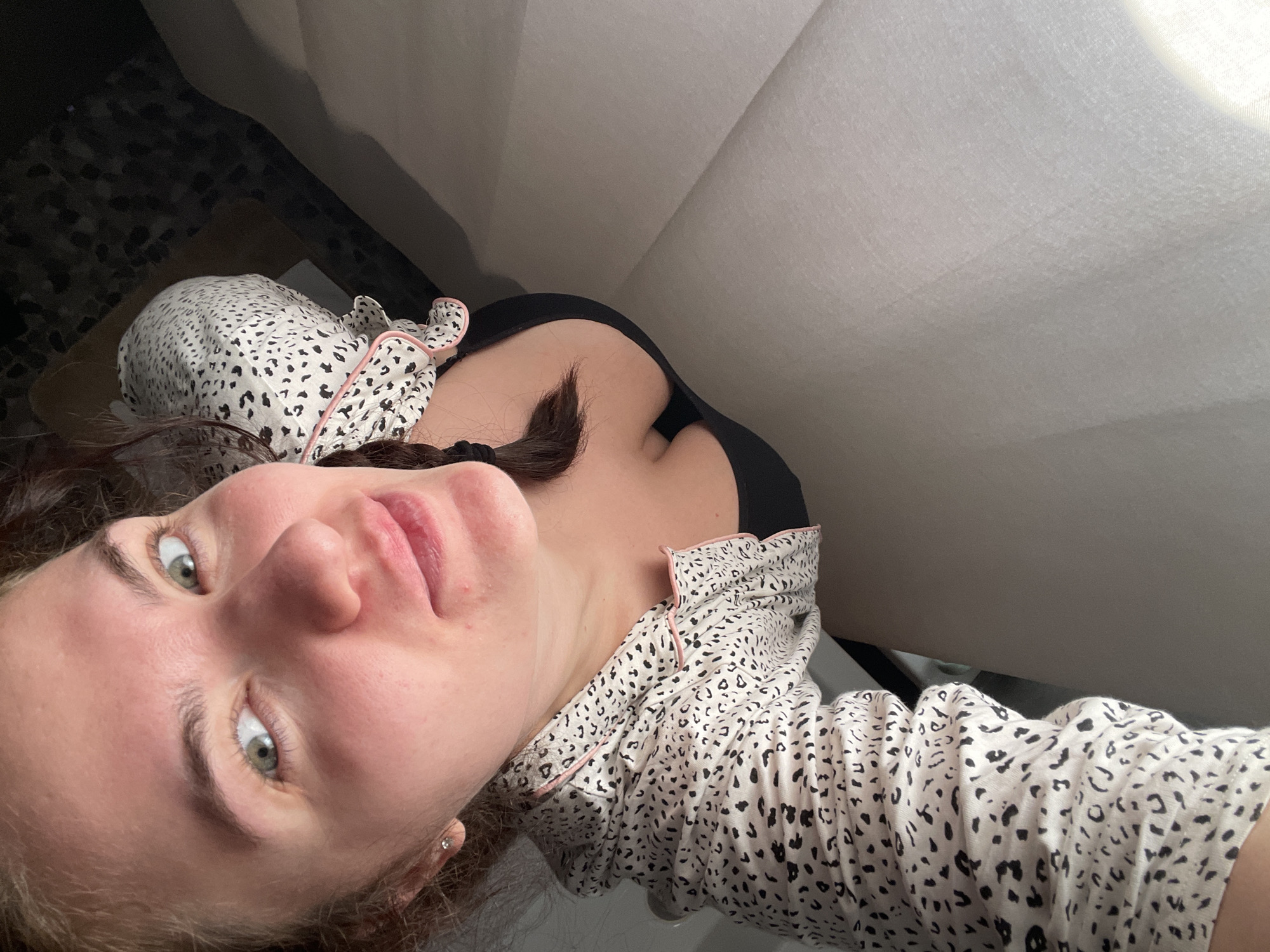 A woman lies down, close to the camera, in a brightly lit indoor space. She is wearing a black bra under a pajama top with a white base and black leopard print with pink trim. A white sofa is in the background to the right. The location is not clearly discernable from the image.