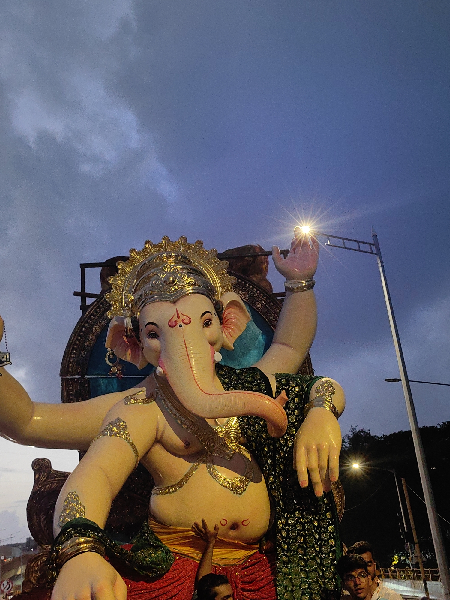 An idol of Lord Ganesha is being paraded through a street in Mumbai, India. The idol is large and adorned with a golden crown and jewelry. It is dressed in a green and gold outfit. The sky above is dark and cloudy, suggesting it might be evening or nighttime. A street lamp with a bright light is visible in the background, as are other people who are part of the procession, some looking up at the idol. The scene appears to be part of a festival or religious event.