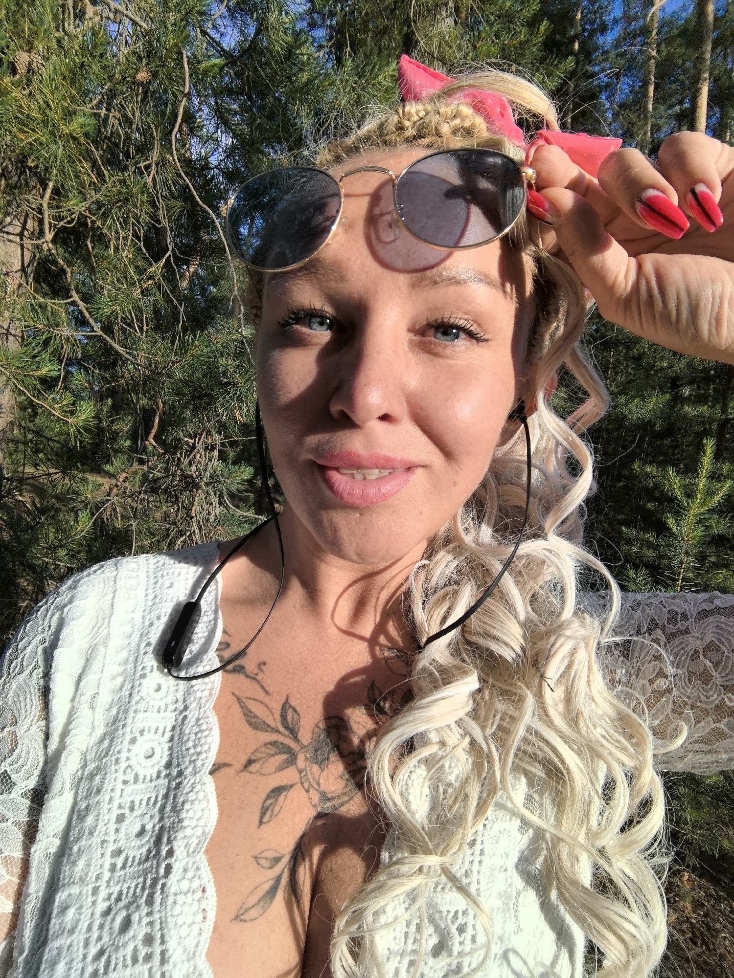 A young woman with blonde curly hair and blue eyes is posing for a selfie outdoors. She is wearing sunglasses and a white lace top. There are tattoos on her chest. She is wearing red nail polish and has a pink ribbon in her hair. The background is a forest with pine trees. The image is taken in Noviy, Russia.