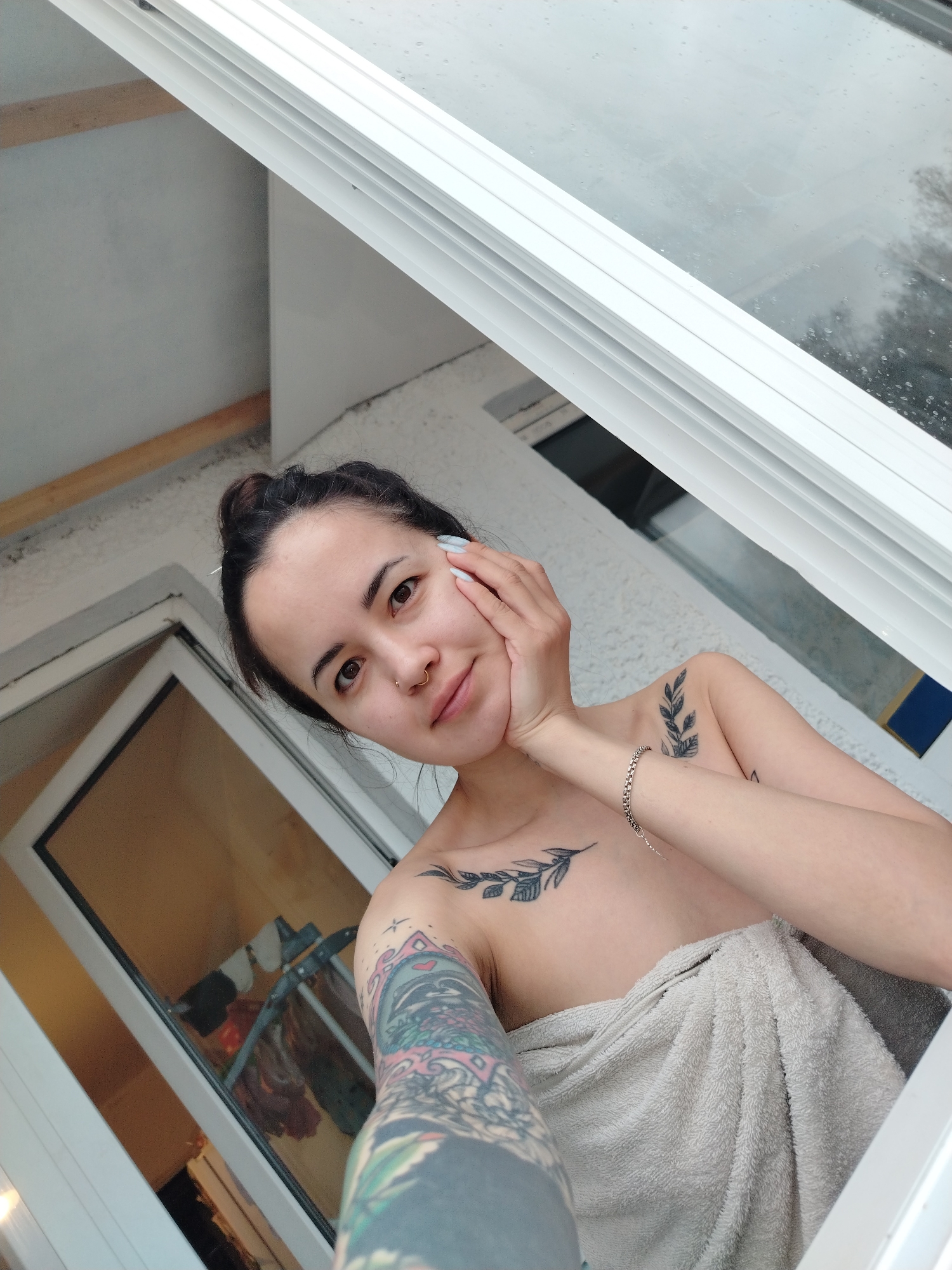 A young woman is featured in a close-up selfie, taken from a slightly low angle, set within what appears to be a domestic space in Noviy, Russia. She has dark hair tied back in a bun and a small hoop nose ring. Her expression is calm and direct, with a subtle smile. Her left arm, extended to take the photo, reveals a colorful, intricate full-sleeve tattoo. Other visible tattoos include a dark leafy branch design on her left collarbone area and another similar design on her right shoulder. She is wrapped in a light beige or grey towel, suggesting she has recently showered or is about to.
Her right hand gently touches her face, showcasing a thin silver-colored chain bracelet on her wrist. The background features prominent white window or door frames, with a large pane of glass above her showing what appears to be an overcast sky with visible water droplets, indicating rainy or damp weather outside. Through a lower glass panel, possibly a door or another window, a reflection of an indoor space is visible, including a clothes drying rack with garments hanging on it. A horizontal wooden beam is also visible in the upper left background, part of the building's structure. The lighting is soft and diffused, consistent with an overcast daytime. No text is visible in the image.
