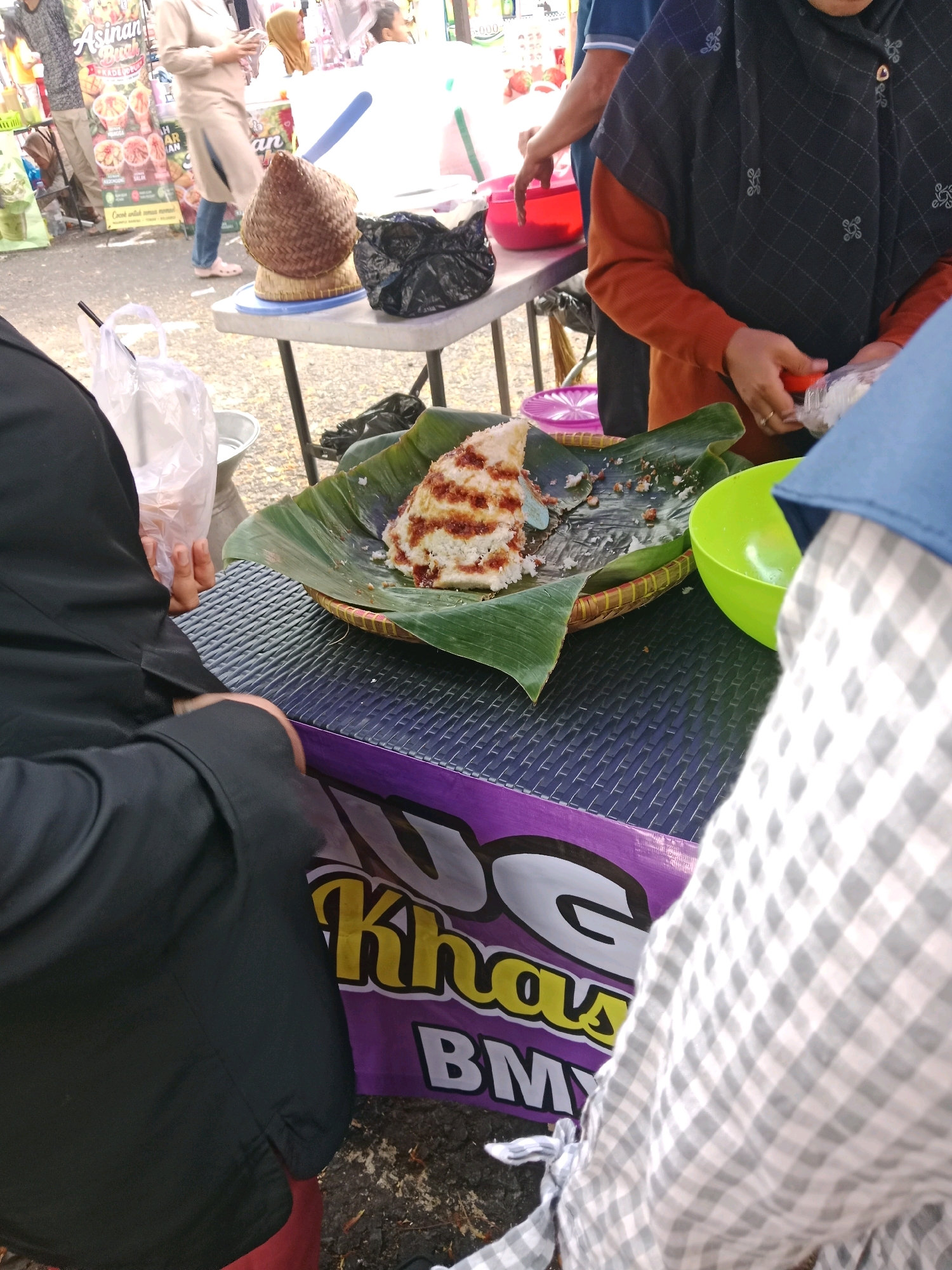 The image depicts a street food stall likely located in Ciamis, Indonesia, given the presence of Indonesian text on signage. A woman in a black hijab and orange-sleeved top is seen serving food, holding a small red lid. In front of her, on a woven mat lined with banana leaves, is a portion of a layered rice cake with a reddish topping, suggesting a sweet or savory dish. A green bowl is positioned to the right of the food, and a clear plastic bag with a straw is on the table to the left. The table is covered with a dark, textured mat, and a purple banner with yellow text that appears to be in Indonesian, possibly reading "Kha" and "BMY," is visible in the foreground. In the background, other individuals are present, with one person wearing a beige top and blue jeans and another in a black and grey patterned top, suggesting a bustling market or outdoor event. The ground is paved with asphalt, and scattered leaves are visible. The overall scene captures a moment of food preparation and transaction at a local Indonesian food stall.