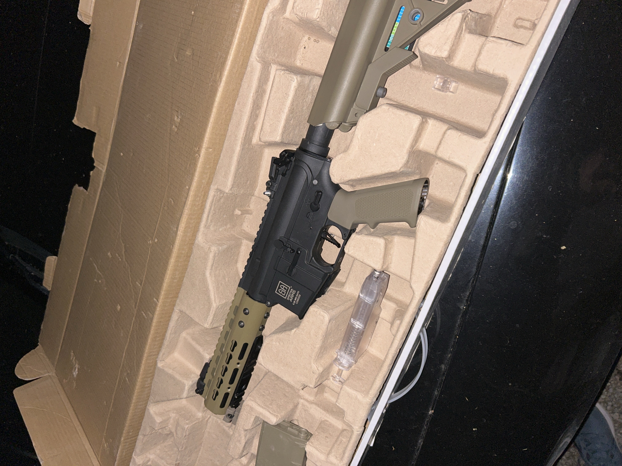 The image shows an airsoft rifle inside its cardboard packaging, placed on a black surface. The rifle has a black receiver, an olive green stock, handguard, and pistol grip. A transparent magazine and an olive green magazine are also visible inside the packaging. The cardboard packaging is brown and provides molded cushioning for the rifle. The image does not contain sufficient information to provide a location in the city of Veliko Tarnovo, Bulgaria.