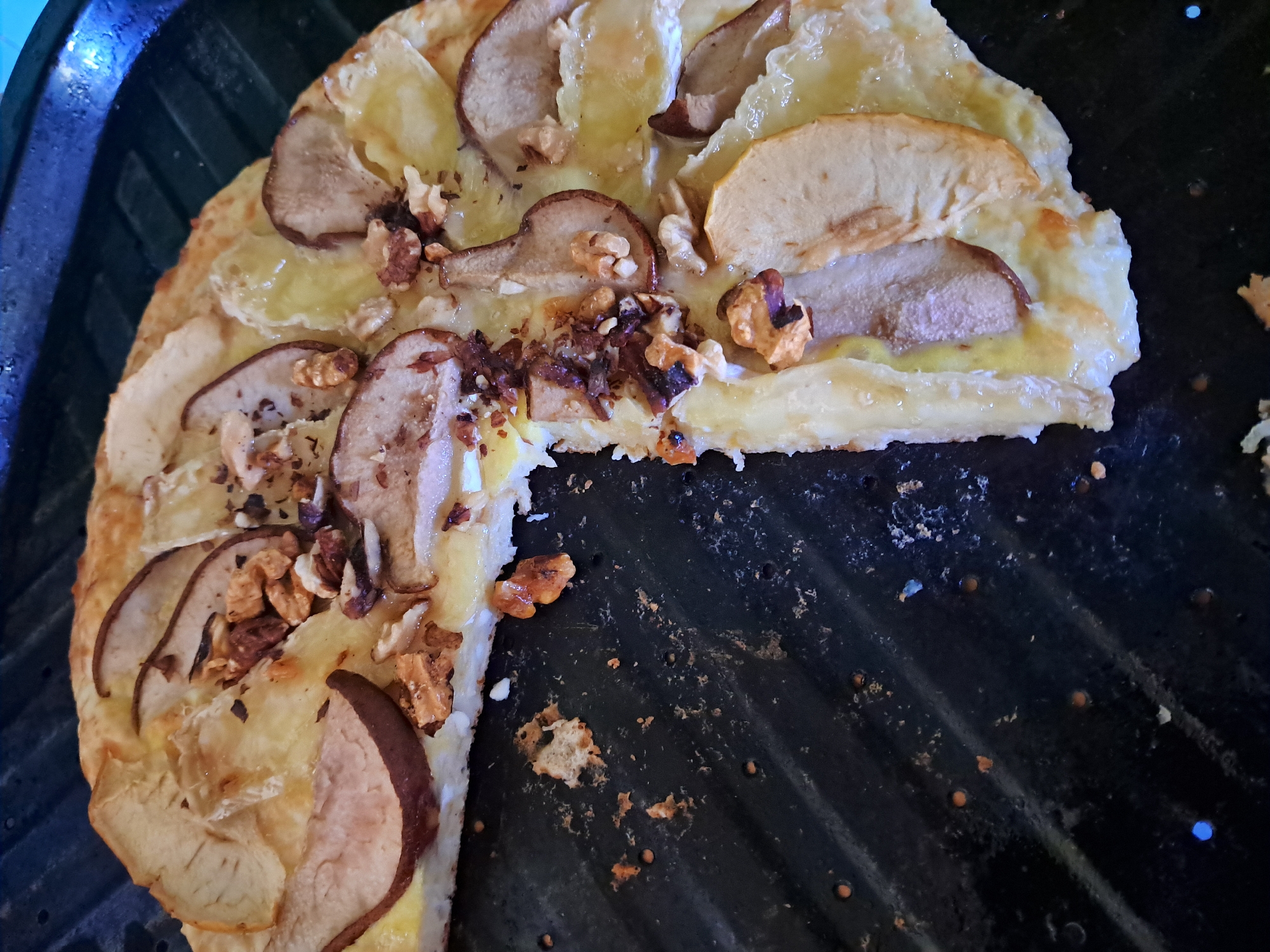 A close-up, overhead shot shows a partially eaten pizza with thin crust, topped with melted cheese, thinly sliced pears, and chopped walnuts, set against a dark, ribbed surface. The pizza has a light golden crust and the toppings are scattered unevenly. Some pieces of crust and toppings have fallen onto the dark surface, which appears to be a grill pan. The lighting is somewhat dim and casts shadows, highlighting the texture of the food and the pan. There is no indication of people or location in the city of Unknown.