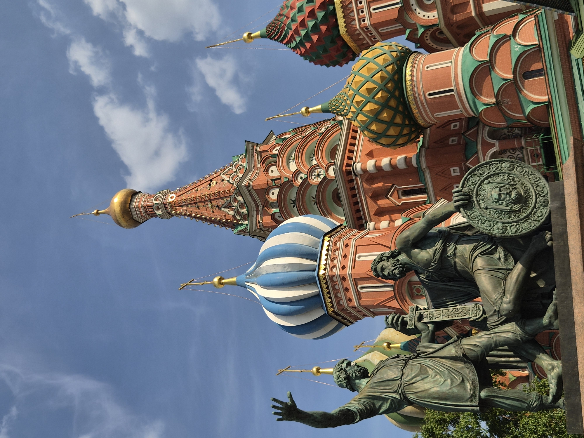 The image displays the Monument to Minin and Pozharsky positioned in the foreground, with Saint Basil's Cathedral in the background, set against a bright blue sky with sparse white clouds. This scene is located in Red Square, Moscow, Russia.
The monument, rendered in bronze, features two male figures. The figure on the left stands, extending his right arm upward with an open palm, and is clad in a tunic and boots. The figure on the right sits, holding a sword in his right hand and a circular shield featuring a sculpted face in his left. Both figures have beards and appear to be in a dynamic pose. Green foliage is visible behind the standing figure's lower body on the left.
Saint Basil's Cathedral occupies the mid-ground and background, showcasing multiple colorful onion domes and spires. A tall, central spire with a golden dome and a reddish, textured conical roof dominates the upper-center. To its left, a large dome is striped vertically in blue and white. To the right, another dome is patterned in green and yellow, and a smaller one beyond it is patterned in red and green. Each dome is topped with a golden finial and an Orthodox cross. The cathedral's main structure is constructed from red brick, accented with white trim and green roofing elements. The architecture features multiple arches, windows, and decorative cornices. The lighting indicates daylight conditions.