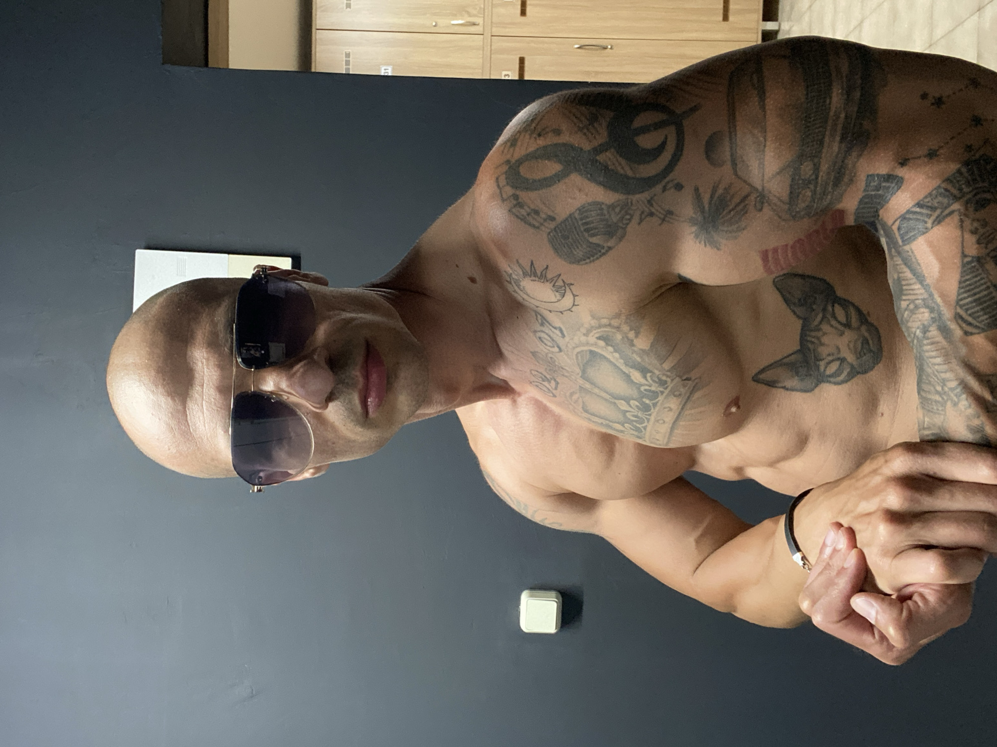 A bald man with sunglasses and tattoos is posing shirtless in front of lockers. The scene is set in a gym locker room. The man has muscular arms and chest. He is wearing dark sunglasses. His tattoos are visible on his chest, arms, and torso. The lockers are made of light-colored wood and are stacked in two rows. There is a light switch on the wall behind him. The overall impression is one of a fitness enthusiast in a locker room environment.