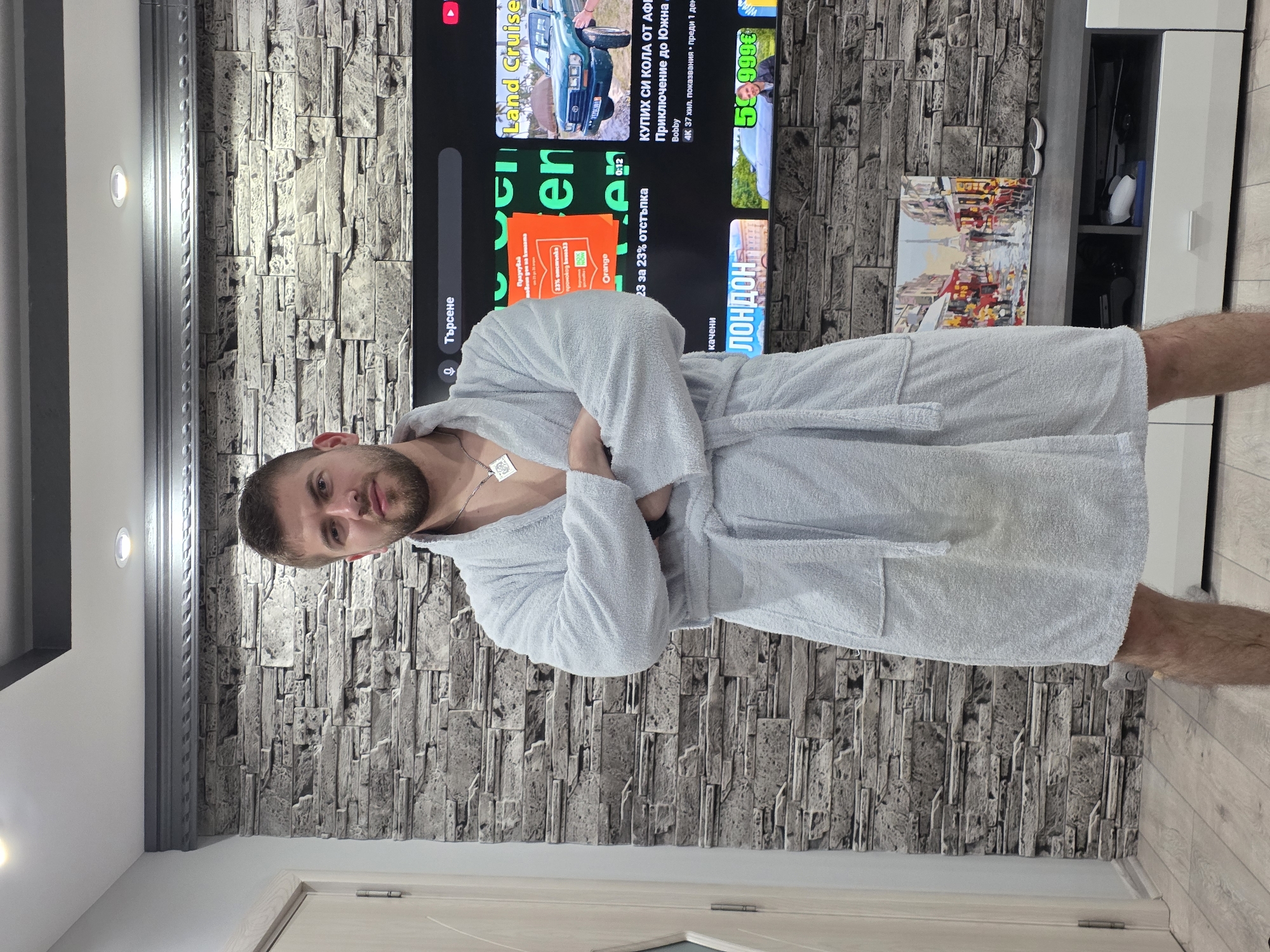 In an indoor setting in Yagodovo, Bulgaria, a man is captured standing and posing for the camera. He appears to be in his 20s or 30s, with a beard, and is wearing a light-colored, possibly light blue or grey, terrycloth bathrobe, with his arms crossed over his chest. A silver necklace with a rectangular pendant hangs around his neck. He maintains a neutral to slightly serious expression while looking directly forward.
Behind him, a large television screen is visible, displaying a YouTube-like interface with various video thumbnails and text, predominantly in Bulgarian. The wall behind the TV features decorative grey cladding resembling stacked stone or brick. On the screen, visible text includes "Търсене" (Search), "Land Cruise" associated with an image of a green off-road vehicle, and an advertisement for "Orange" with text such as "Празнувай 23% отстъпка промокод вина33" (Celebrate 23% discount promo code wine33) and "23 за 23% отстъпка" (23 for 23% discount). Another video title reads "КУПИХ СИ КОЛА ОТ АФР Приключение до Южна Bobby" (I BOUGHT A CAR FROM AFR Adventure to Southern Bobby), noted as "4К 37 хил. показвания преди 1 ден" (4K 37k views 1 day ago). The word "ЛОНДОН" (LONDON) is also visible near a thumbnail, along with a price "59999€" and the word "качени" (uploaded/added).
To the right of the TV, a white cabinet with open shelving holds various items. A colorful framed painting of a cityscape, possibly Paris, rests on a surface below the TV. The flooring is a light-colored wood or laminate. Recessed lighting fixtures and a decorative ceiling molding are visible above. The scene depicts a contemporary home interior.