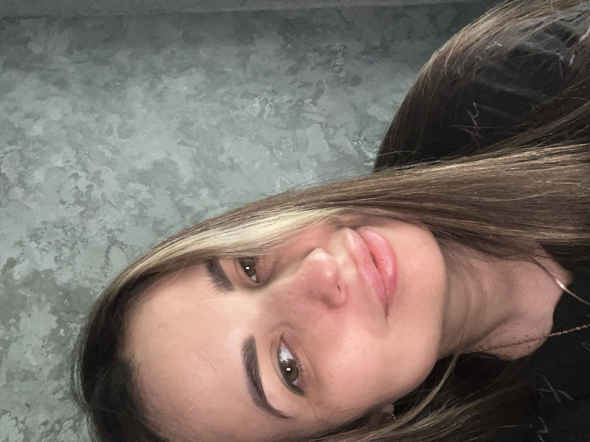 A close-up, slightly angled shot of a woman's face. She has light skin, brown hair with blonde highlights, and brown eyes. Her eyebrows are well-defined. She is wearing a dark-colored shirt. A thin gold necklace is visible around her neck. The background appears to be a textured gray wall.
Location: Unknown, Unknown.