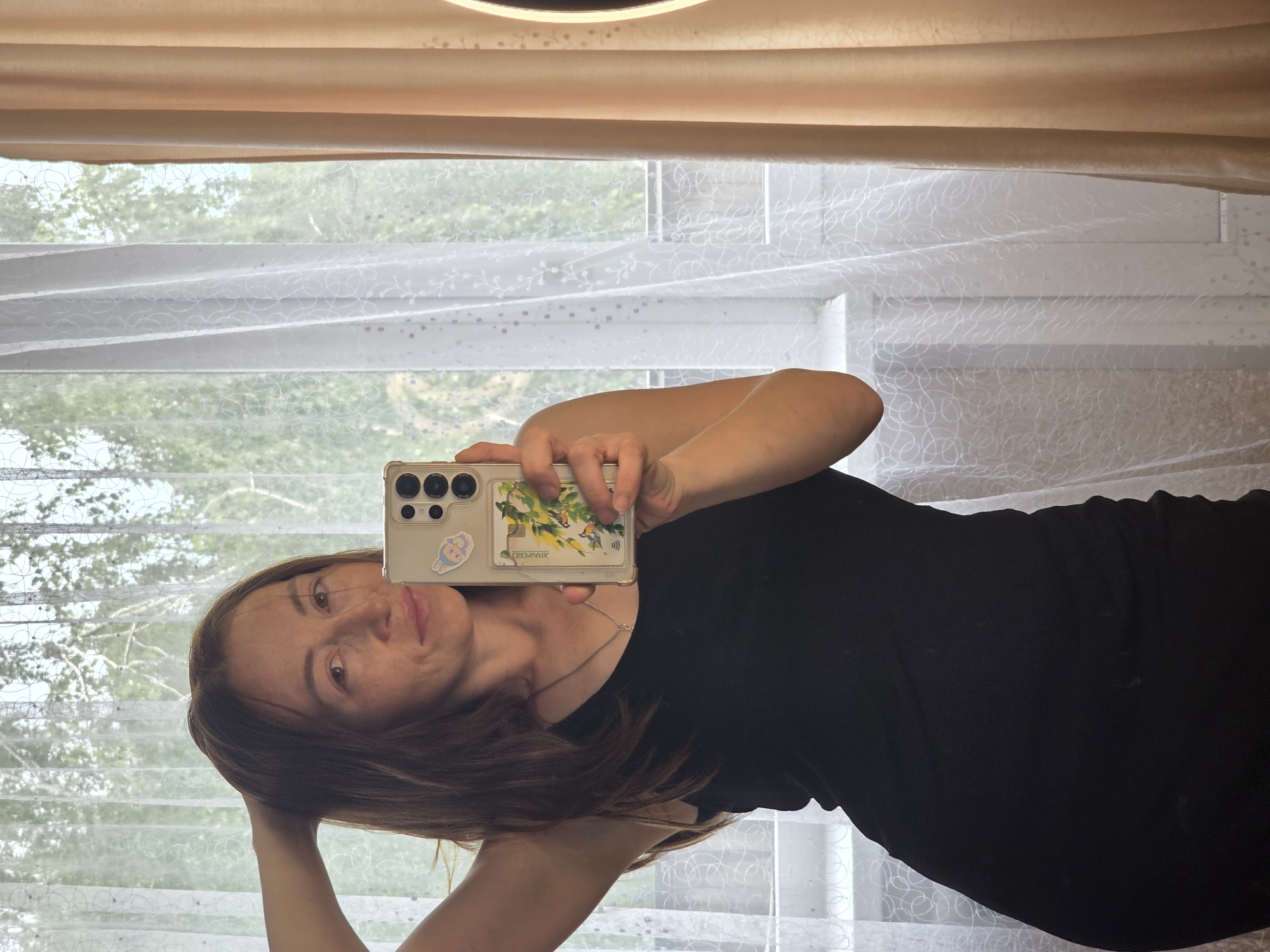 A woman with long, brown hair is taking a mirror selfie. She is wearing a black tank top and a silver necklace. Her right arm is raised and her hand is in her hair. She is holding a smartphone in her left hand, which is in front of her chest. The phone has a case with a sticker of a bunny and a card holder with an image of birds and branches. The background appears to be a window with white sheer curtains and a beige curtain to the right. Sunlight is filtering through the curtains, illuminating the scene. The environment suggests an indoor setting, possibly a home.