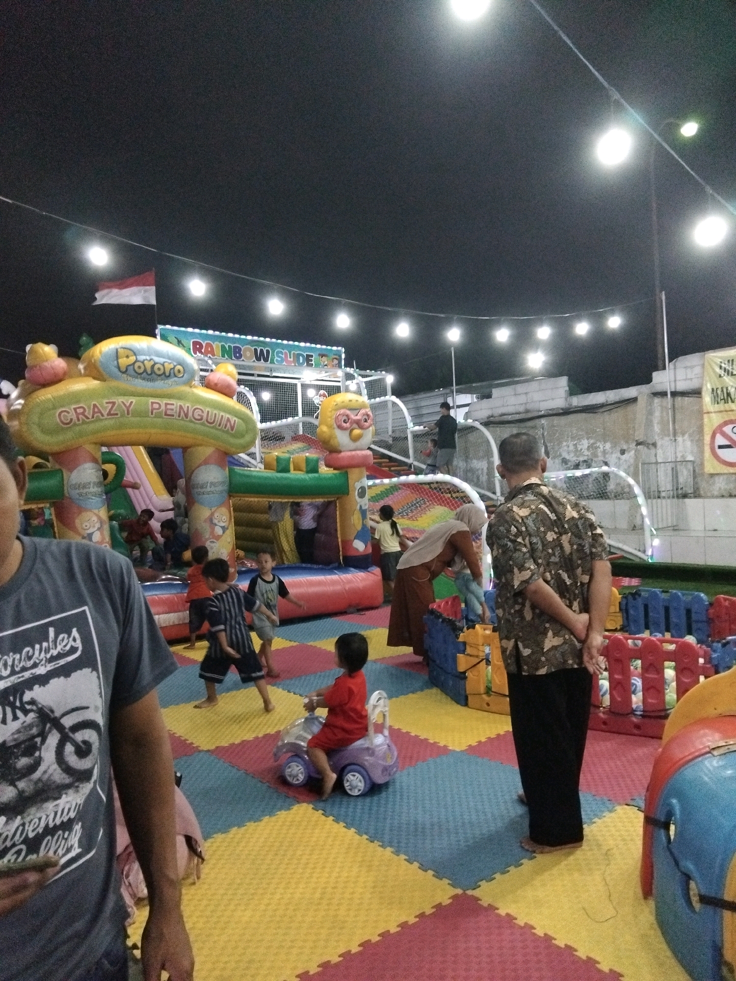 In Sedati Agung, Indonesia, an outdoor amusement park is bustling with activity at night. The park features an inflatable playground with a "Crazy Penguin" theme and a "Rainbow Slide." Several children are engaged in play, including a child riding a toy car and others navigating the inflatable structures. Adults are present, supervising and interacting with the children. The environment is illuminated by string lights and spotlights, casting a warm glow on the colorful play area. The ground is covered with a padded matting in a checkered pattern of yellow, blue, and red. A partially visible person in the foreground is wearing a grey t-shirt with a motorcycle graphic. In the background, a building structure and a sign with Indonesian text are visible. A red and white Indonesian flag waves in the breeze.