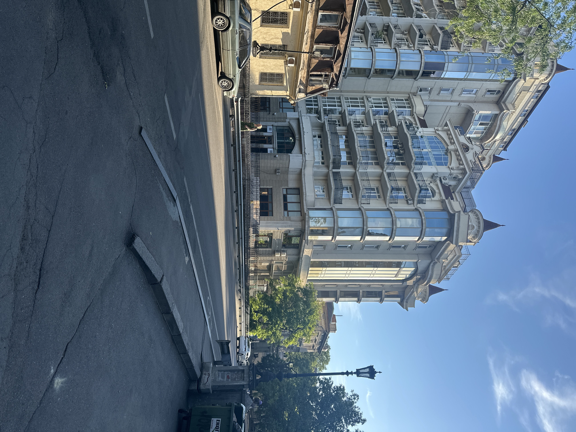 The image shows a street scene in Odesa, Ukraine, on a clear, sunny day. A large, modern residential building with rounded glass balconies and conical turrets dominates the background. To the left of this building is an older, yellowish building with a mansard roof and several windows, one of which is open. A light-colored sedan is parked on the left side of the street, with its front wheels on the sidewalk. A person in a green outfit is walking on the sidewalk in front of the modern building. A black, ornate lamppost stands on the right side of the street, and a green dumpster with Cyrillic writing is partially visible next to it. The street itself is asphalt, with painted lane markings and a curb. Trees line the right side of the street, behind the lamppost.