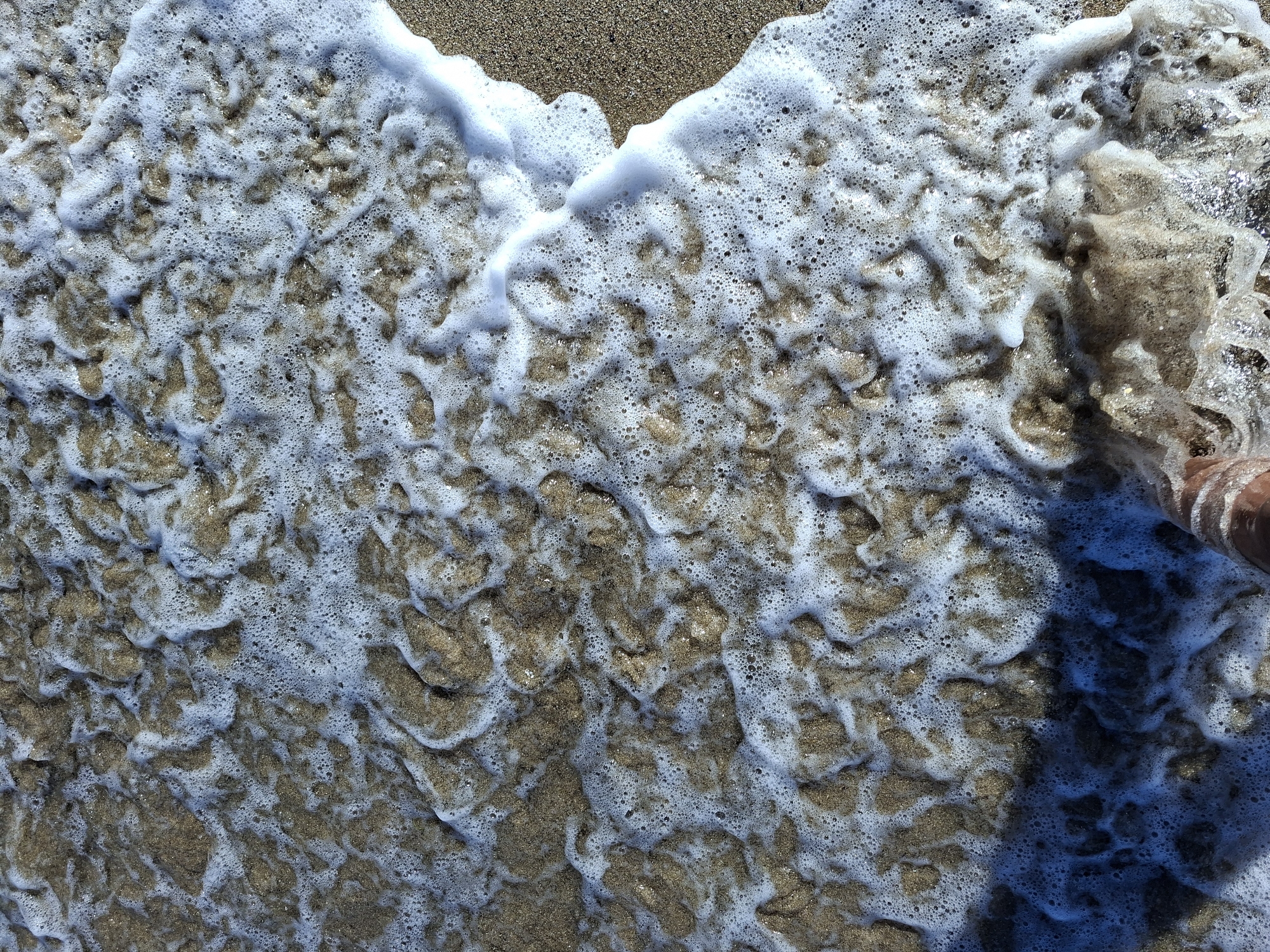 The image is an overhead, close-up shot of a sandy beach with foamy waves rolling in. The sand is a light brown color, and the water is clear with white foam creating intricate patterns as it recedes. A single foot, partially submerged in the water and foam, is visible in the bottom left corner of the frame. The lighting suggests it is daytime, with sunlight reflecting off the water and sand. There are no buildings or specific city landmarks visible, making it impossible to determine a specific location within a city.