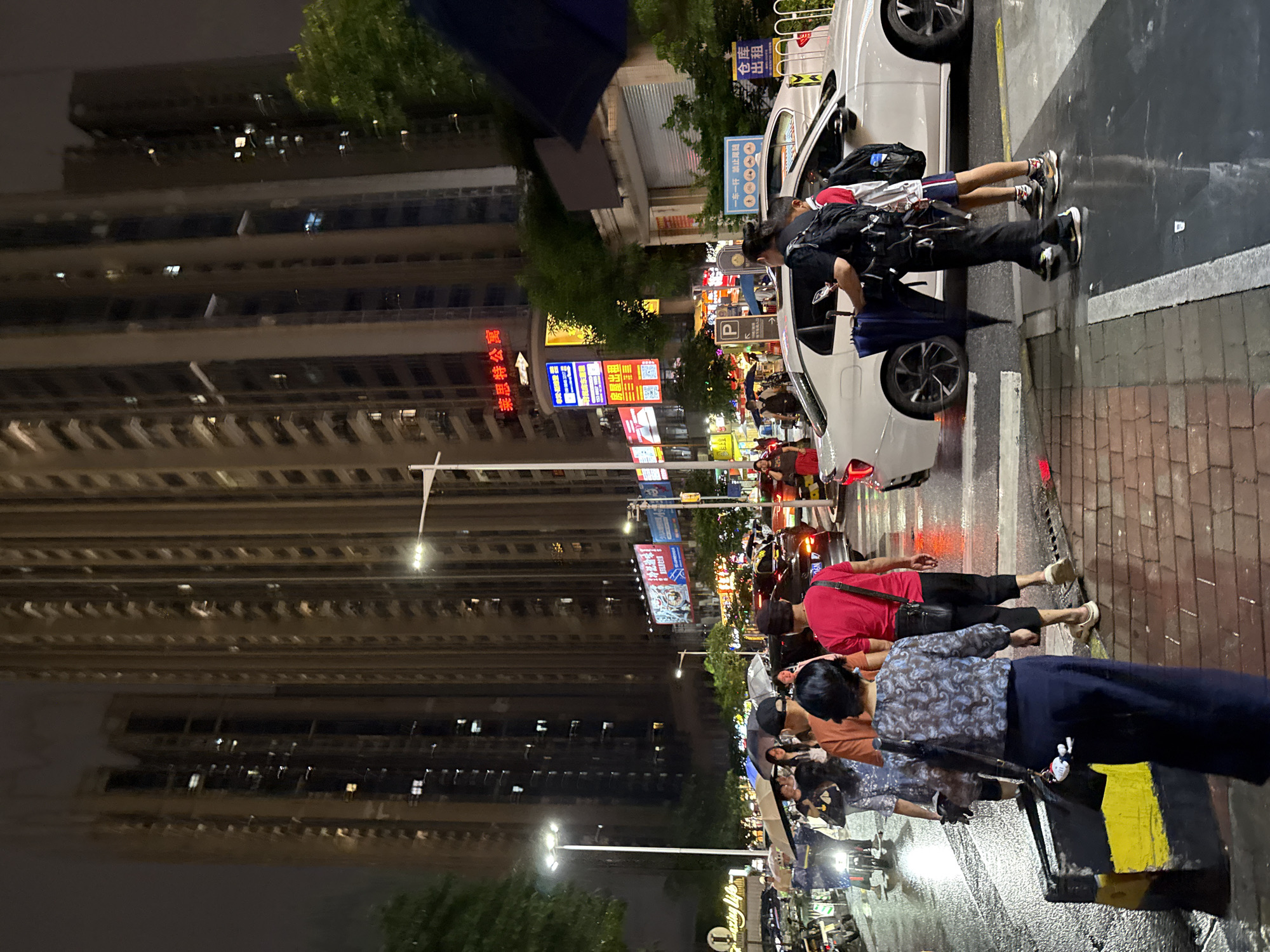 This image captures a bustling street scene at night in Haizhu District, China. The foreground is dominated by a white car stopped at a pedestrian crossing, with several people gathered around it. One individual, dressed in black, is leaning into the car, while another, a young person in a red shirt and shorts, stands beside them. The ground is wet, suggesting it has recently rained, and the streetlights reflect off the slick surface.
In the background, tall, illuminated apartment buildings rise into the night sky. The buildings are modern, with many windows showing lights from within, indicating a residential area. A variety of signs with Chinese characters and some English text are visible along the street, including advertisements for businesses and directional information. One sign reads "莱思特公寓" which translates to "Leicester Apartment". Another appears to be a parking sign with a "P" logo and the text "禁止尾随", meaning "No Tailgating".
The overall atmosphere is one of urban nighttime activity. The presence of multiple people, parked cars, and commercial signage points to a lively commercial or residential area. The lighting from the signs and buildings creates a vibrant, almost neon-like glow, contrasting with the darker sky. Despite the wet ground, the scene doesn't convey any sense of distress, rather a typical evening in a populated city. The text "Amazing Life" is also visible, suggesting a positive or aspirational marketing message.