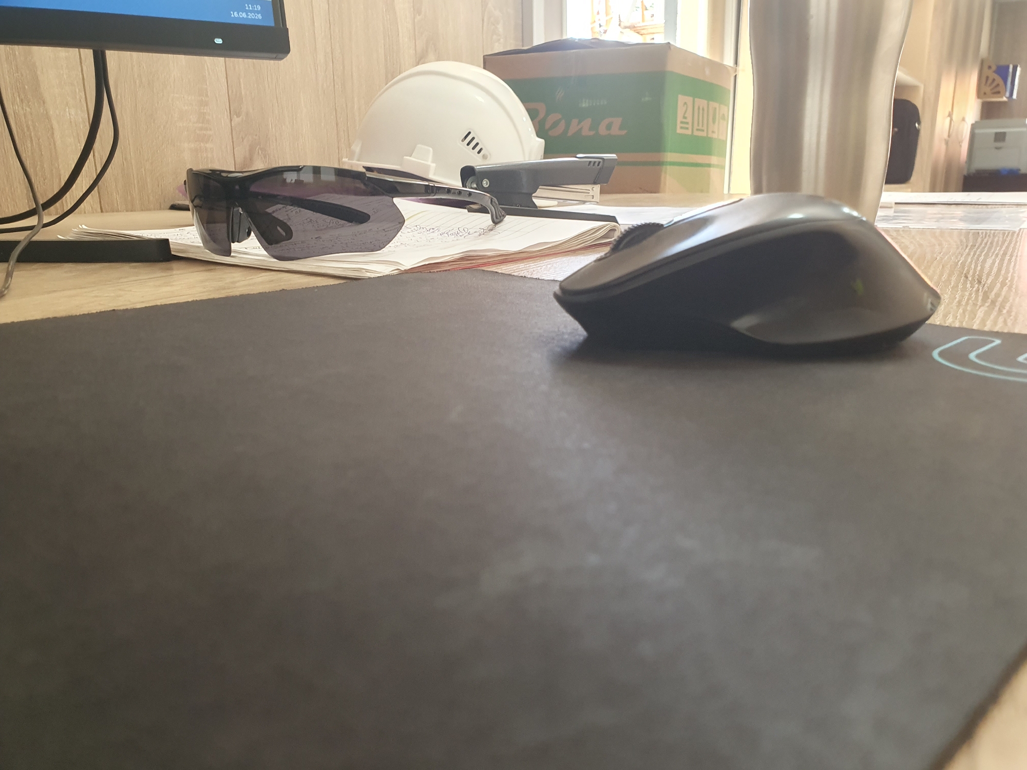 The image shows a desk with various items, including a computer monitor, a hard hat, safety glasses, a stapler, a notebook, and a computer mouse. The scene appears to be in an office or workshop environment, with wooden furniture and a window in the background. There are no people visible in the image. The location is the city of Tashkent, Uzbekistan.