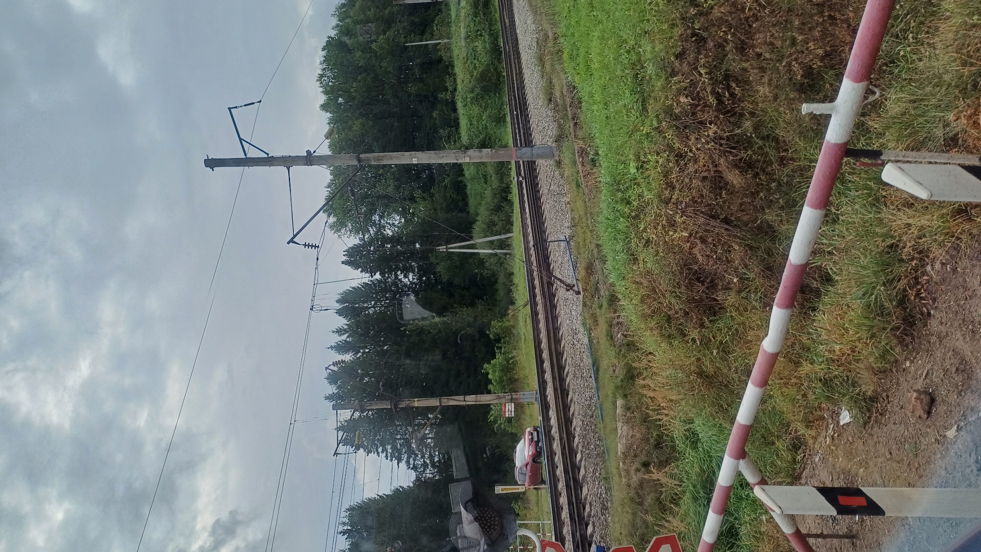 The image depicts a railroad crossing in a rural setting, likely in or near the village of Kechevo, Russia, given the provided context. A red car is stopped at the crossing, positioned on the right side of the frame near the tracks. Railroad tracks extend horizontally across the mid-ground, with ballast and a concrete barrier visible. Overhead power lines for the train are supported by wooden poles. The surrounding environment is characterized by dense green trees forming a forest in the background and tall grass and brush in the foreground. A red and white striped railway crossing barrier is prominently displayed in the lower portion of the image, with a warning sign attached to its post. The sky is overcast and cloudy. The overall scene suggests a typical day at a rural railway crossing.
