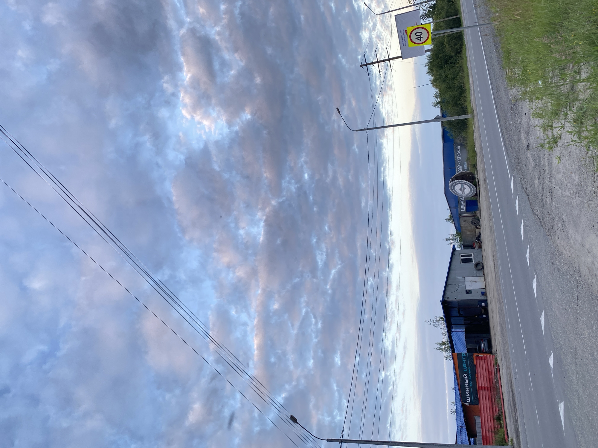The image depicts a scene in the city of Nyagan, Russia, with a road in the foreground and a commercial building on the right side of the road. The sky is filled with clouds, indicating it is likely dusk or dawn.
In the foreground, a paved road runs diagonally across the image from the bottom left to the upper right. White triangular markings are visible on the road, suggesting a traffic lane. To the right of the road, there is a grassy area with some sparse vegetation.
On the right side of the road, a speed limit sign indicating "40" is prominently displayed on a yellow background, mounted on a pole. Above the speed limit sign, there is a larger sign with text that appears to be related to elections. Further down the road, a utility pole with power lines is visible.
In the middle ground, a long, low-slung building complex runs along the side of the road. The building has a blue roof and appears to be a commercial establishment, possibly a tire shop or service center, as suggested by a banner with text that reads "ШИННЫЙ ЦЕНТР" (Tire Center) and "ЗАПАДНЫЙ" (Western). There is a red fence in front of part of the building on the left. A car is parked near the building. Several large tires are stacked outside the building.
The background is dominated by the sky, which features a mix of blue and gray clouds, with some patches of light suggesting the sun is either rising or setting. The overall impression is that of a somewhat industrial or commercial area on the outskirts of a city under a dramatic sky.
