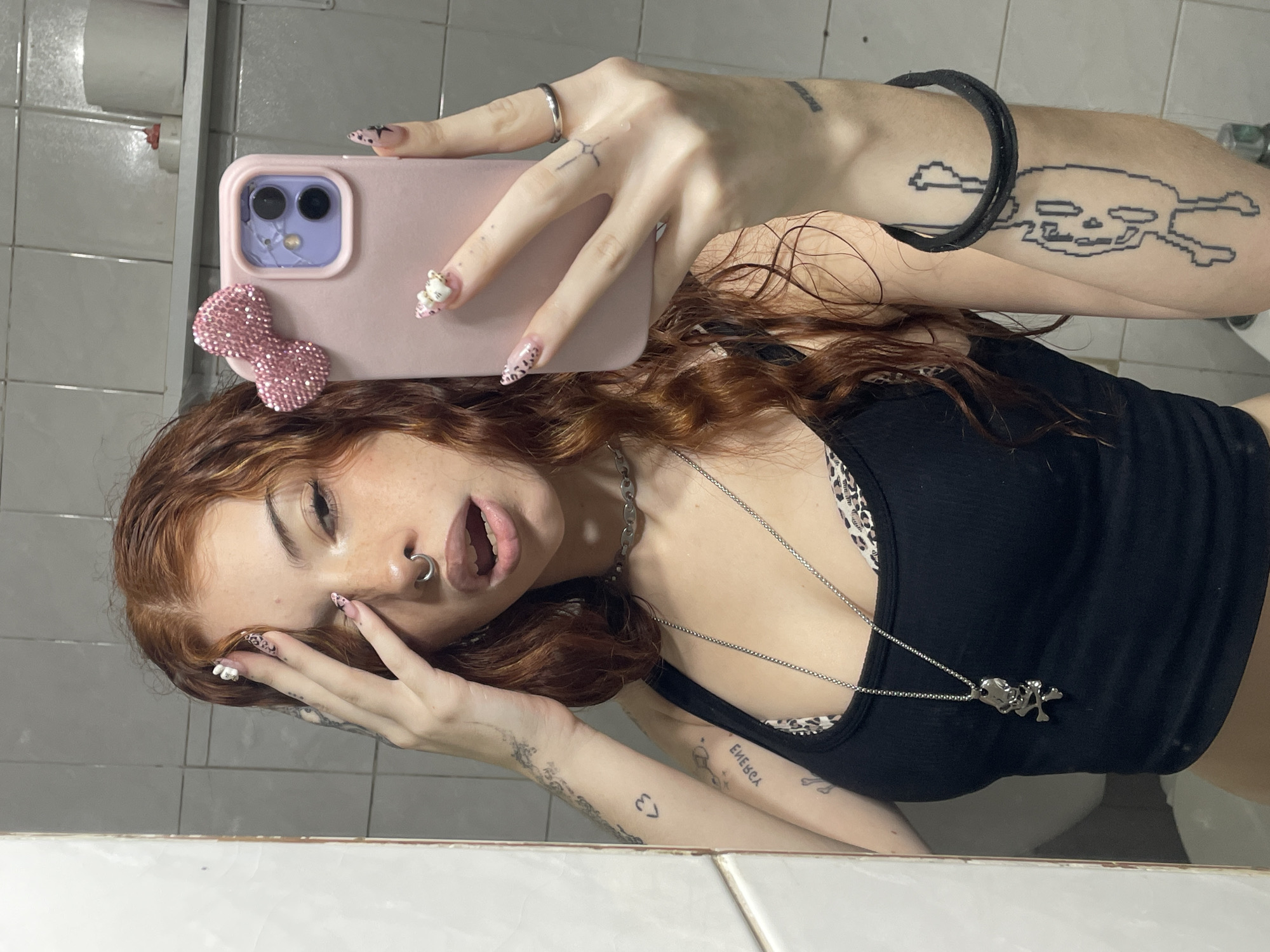 A mirror selfie captures a young individual with long, wavy reddish-brown hair, posing in what appears to be a bathroom. The person is wearing a black ribbed tank top over a leopard print bralette, and has a silver hoop nose ring. Their long, pointed nails are decorated with intricate designs, including leopard print and a 3D Hello Kitty charm on one finger.
The individual holds a pink smartphone, adorned with a sparkly pink bow accessory, in their right hand. The phone's camera lenses are purple, with one showing a crack. Various tattoos are visible: a large pixelated skull and crossbones on the right upper arm, a small cross-like symbol on the right hand, and abstract wavy patterns on the left forearm. On the inner left forearm, a small heart symbol '<3' and the reversed text 'ЕМЕВСА' (likely 'ACEBME' when viewed correctly) are tattooed. A silver chain necklace with a heart pendant and another charm hangs around their neck.
The background, reflected in the mirror, consists of light-colored tiled walls and a white surface, with a toilet paper holder visible. The person’s mouth is slightly open, and their left hand is near their face, contributing to a playful and expressive pose.