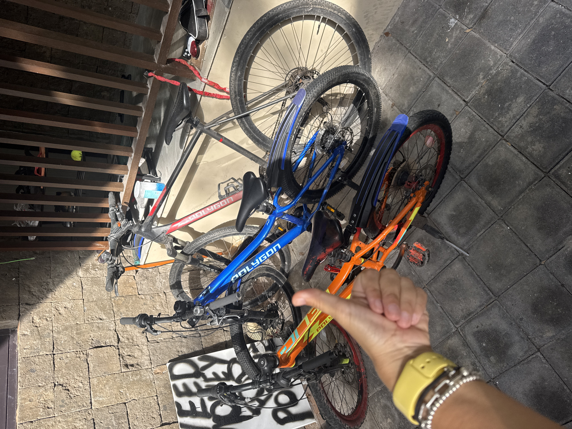 The image shows three Polygon brand bicycles on a tiled surface. The bikes are positioned close together, with a red/gray, a blue, and an orange frame visible. The tires appear to be mountain bike tires. A hand and arm wearing a yellow watch and bracelets are visible in the foreground. The location is Kuta Selatan, Indonesia. There are stairs nearby, casting shadows on the bicycles and the tiled surface.