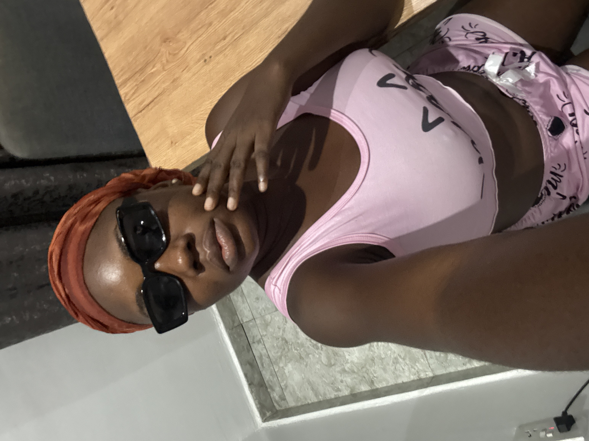 The image captures a person, likely female, with dark skin, in what appears to be an indoor setting. The person is wearing a pink sleeveless crop top adorned with black V-shaped designs, and pink patterned shorts featuring small black cartoon-like graphics and some indecipherable stylized text, with a white drawstring visible. An orange-brown head covering is worn, and black rectangular sunglasses cover the eyes. One hand with manicured nails is raised, gently touching the face near the sunglasses. The person's expression is relaxed, with slightly parted lips.
The background comprises several distinct surfaces: a smooth, light brown wooden surface occupies the upper left, beneath which is a dark gray or black textured surface, possibly part of a couch or chair. The lower right section of the image shows a light gray tiled floor with visible grout lines, and a clean white surface, possibly a wall or fixture, runs along the bottom and right edges of the frame. In the bottom right corner, a white power outlet with a black cord plugged into it is visible. The lighting suggests an indoor environment. The image is taken from a slightly elevated, tilted perspective, indicative of a selfie or a casual portrait. The location is Iddo Tudunwada, Nigeria.