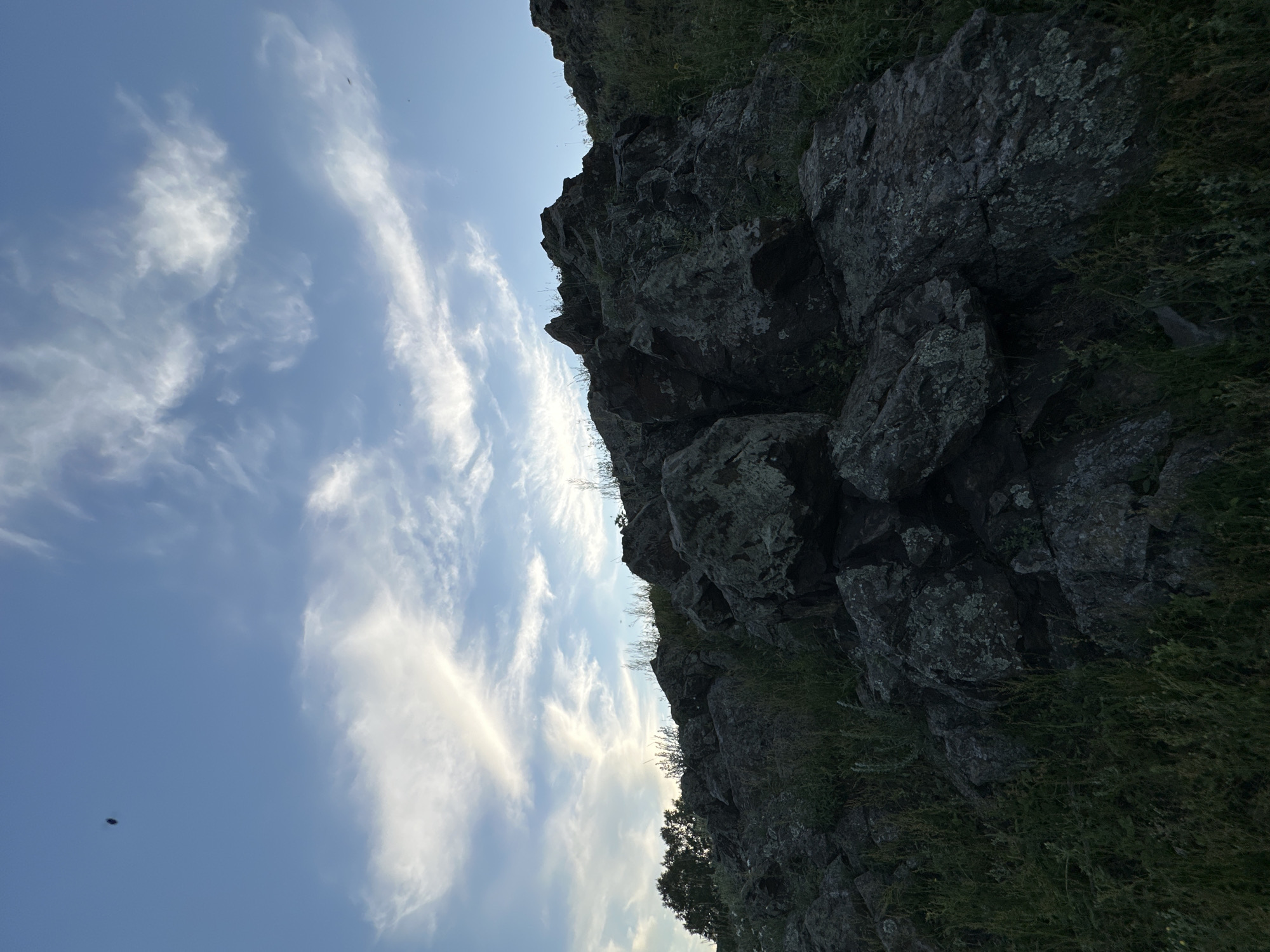 An outdoor, eye-level, wide-angle shot captures a rocky hillside under a partly cloudy blue sky. The foreground is dominated by large, dark grey rocks with textured surfaces, some covered in patches of light green lichen. Green grass and other vegetation grow between and around the rocks, appearing somewhat dry in places. The hillside slopes upwards towards the right of the frame. In the sky, wispy white clouds are scattered across the blue expanse, with a hint of brighter light, possibly from the sun, visible behind some of the clouds on the left. A small, dark, oval-shaped object, possibly an insect or a bird, is visible in the upper left quadrant of the sky. The scene suggests a natural, undeveloped landscape.