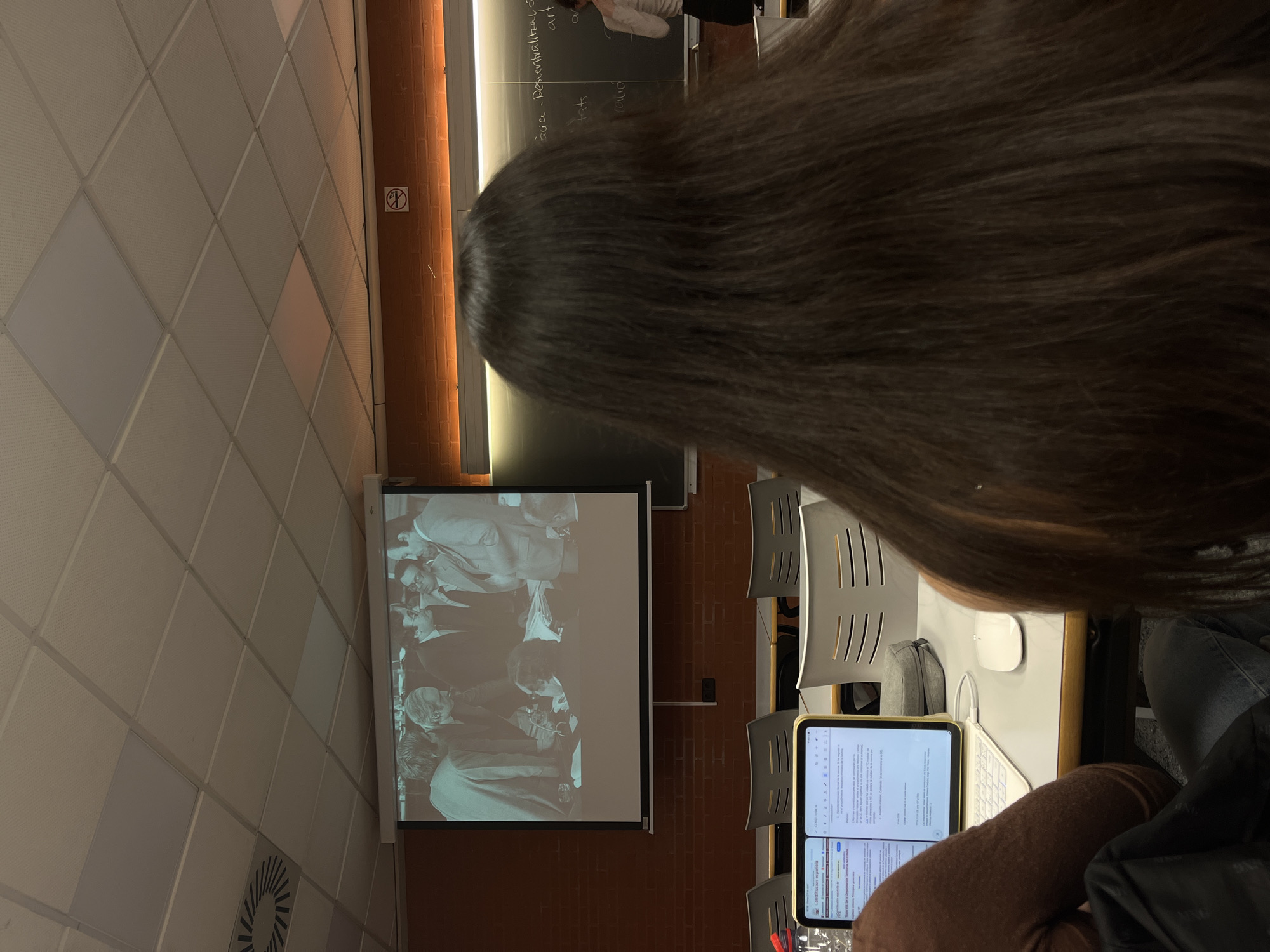 This media file depicts an indoor scene in what appears to be a classroom or lecture hall, likely in Valencia, Spain. The foreground is dominated by the back of a person's head with long, dark hair, suggesting the viewpoint of a seated student.
In front of this person, a wooden desk holds a tablet with a connected white wireless keyboard and mouse. The tablet's screen is split into two panes, displaying text documents. Visible text on the tablet includes "CONSTITUCIÓN ESPAÑOLA" (Spanish Constitution) and "economia política" (political economy), indicating an academic context, possibly related to law or political science. A grey fabric pouch is also on the desk.
To the left, a projector screen displays a black-and-white historical photograph of several men dressed in suits, engaged in what appears to be a formal gathering or discussion. Behind the projector screen, a blackboard is partially visible with handwritten text. Words such as "cària," "Demonialtació," "tats," "art," and "ra...ció" can be discerned, with some terms suggesting Catalan, consistent with the location context of Valencia. A person, possibly a lecturer, is partially visible near the blackboard.
The room's architecture includes a white ceiling with a diamond grid pattern and brown brick walls. Gray, modern chairs are arranged in rows, facing the front of the room. A standard "no smoking" sign is affixed to the wall, and an air vent is visible on the ceiling. The lighting suggests an indoor setting, and the activity appears to be an educational lecture or presentation.