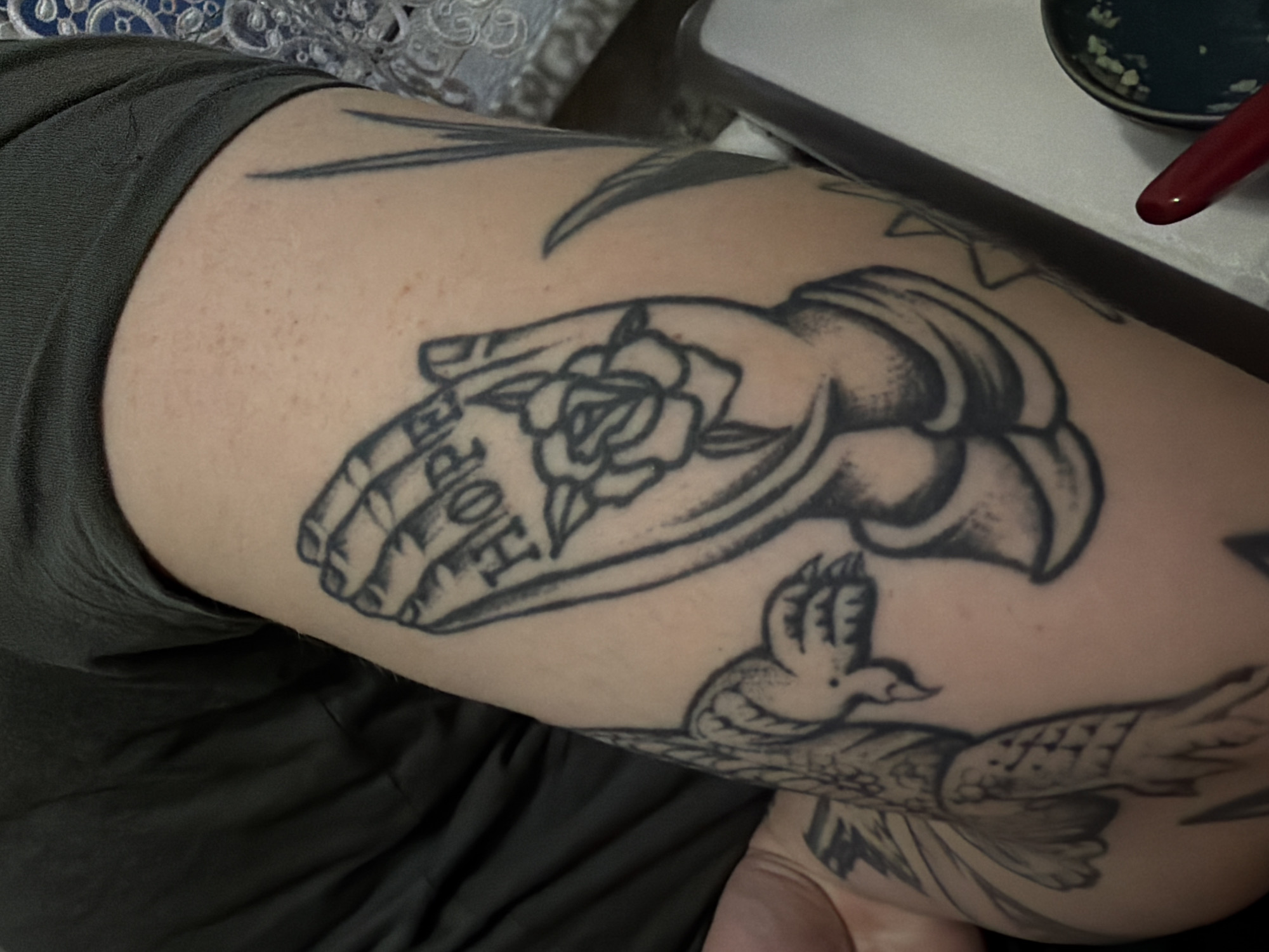 The image shows a close-up of a person's forearm with a black ink tattoo of praying hands. The word "HOPE" is tattooed across the fingers of the praying hands. A rose is depicted within the hands. To the left of the praying hands is a tattoo of a bird, possibly an eagle. The person is wearing a dark green t-shirt. The background is out of focus and appears to be an indoor setting with a textured surface.