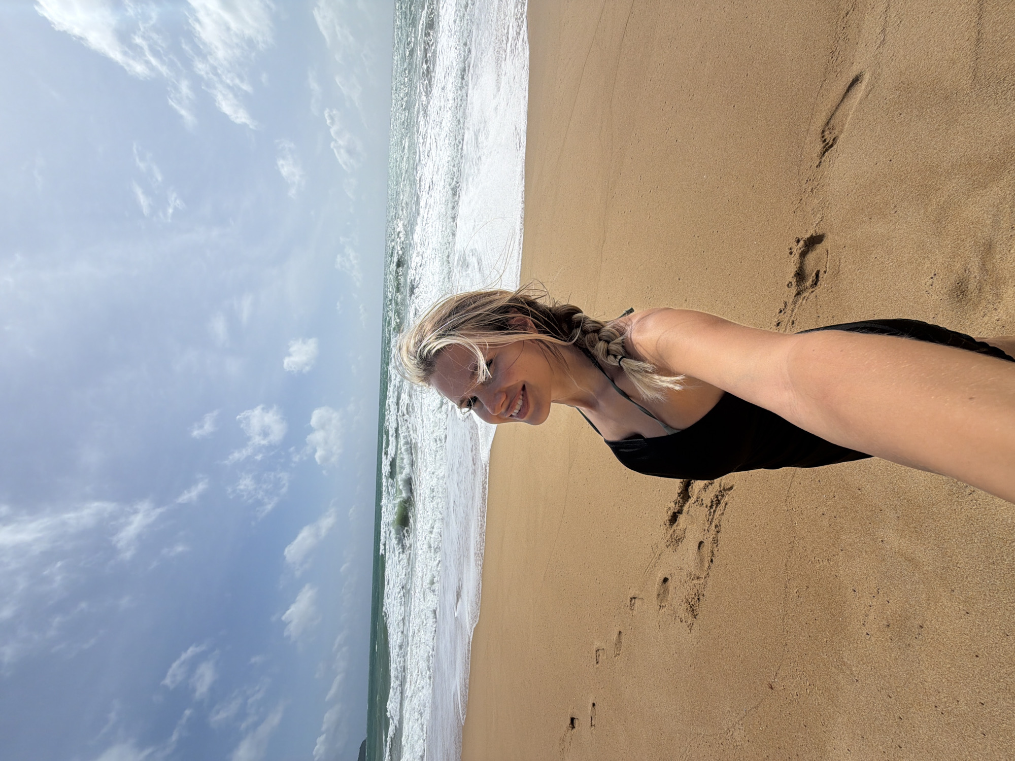 A woman is taking a selfie on a sandy beach with the ocean in the background. She has blonde hair in a braid and is wearing a black swimsuit. The waves are crashing on the shore, and there are footprints in the sand. The sky is blue with some white clouds. The location is Mai Khao Beach, Phuket, Thailand.
