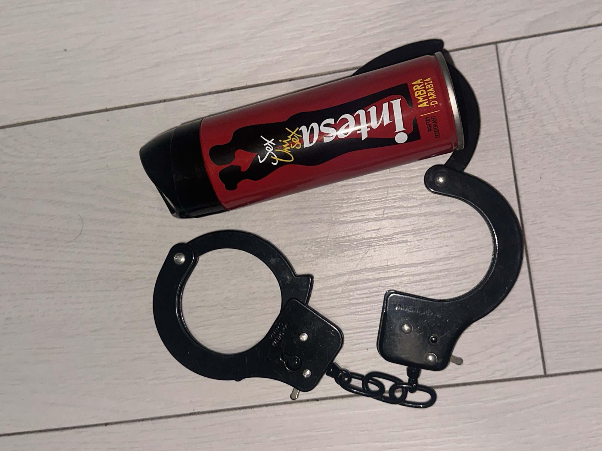 A pair of black metal handcuffs and a red aerosol spray can are positioned on a light-colored, planked floor. The handcuffs are arranged in an open, semi-circular configuration, with one cuff resting near the base of the aerosol can. Text "MADE IN CHINA" is visible on one of the handcuff mechanisms. The red aerosol can features a black silhouette of a man and a woman embracing. White and yellow text on the can reads "Sex Unisex Sex," "Intesa," "PARFUM DEODORANT," and "AMBRA D'ARABIA." The flooring displays faint wood grain and visible seams between planks. The image provides no identifying information regarding its location within the city of Sofia, Bulgaria.