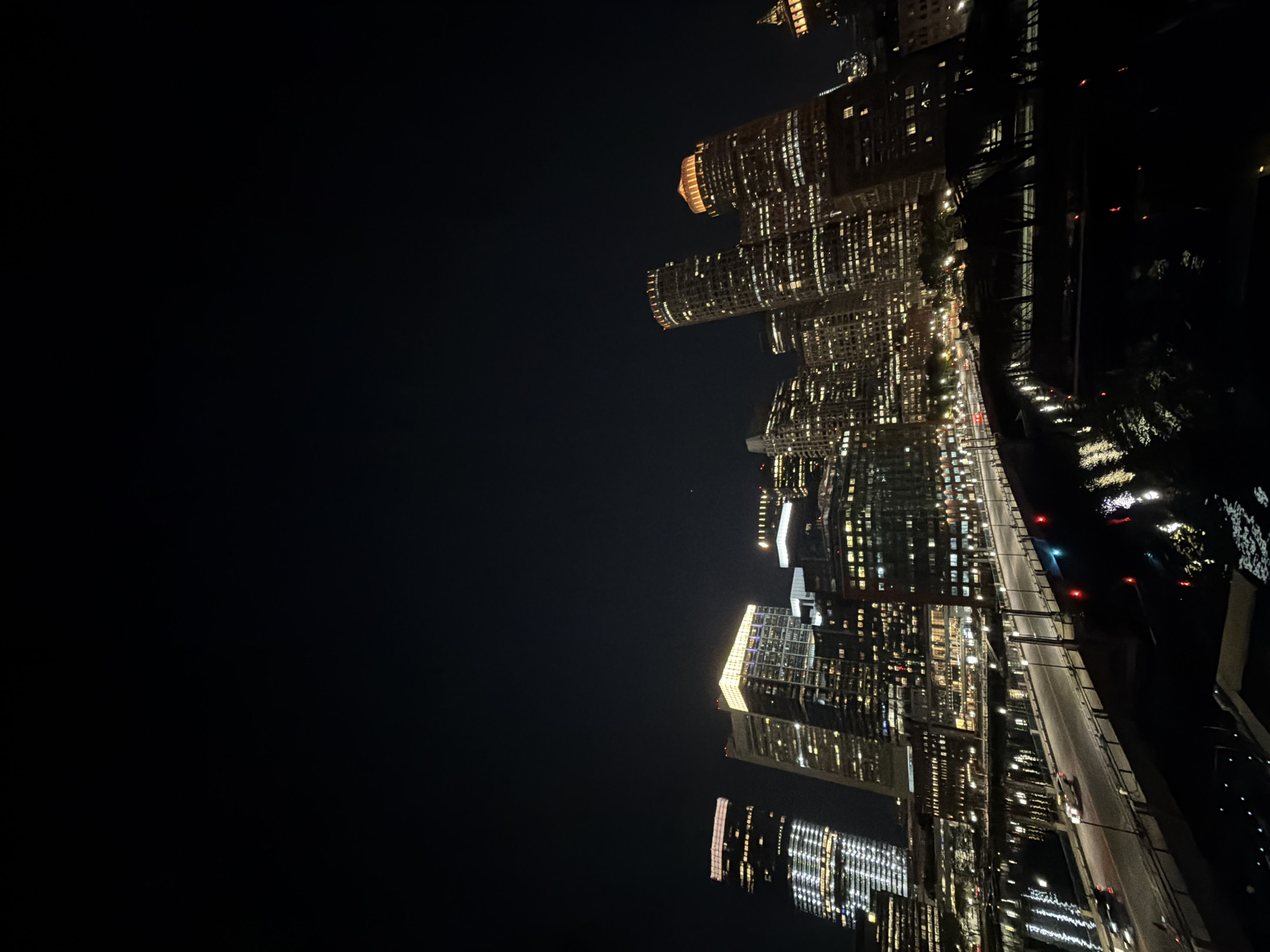 The image shows a nighttime cityscape of Boston, Massachusetts, United States. Prominent skyscrapers line the right side of the frame, illuminated with lights, reflecting on the river. A road runs parallel to the buildings. The sky is dark.