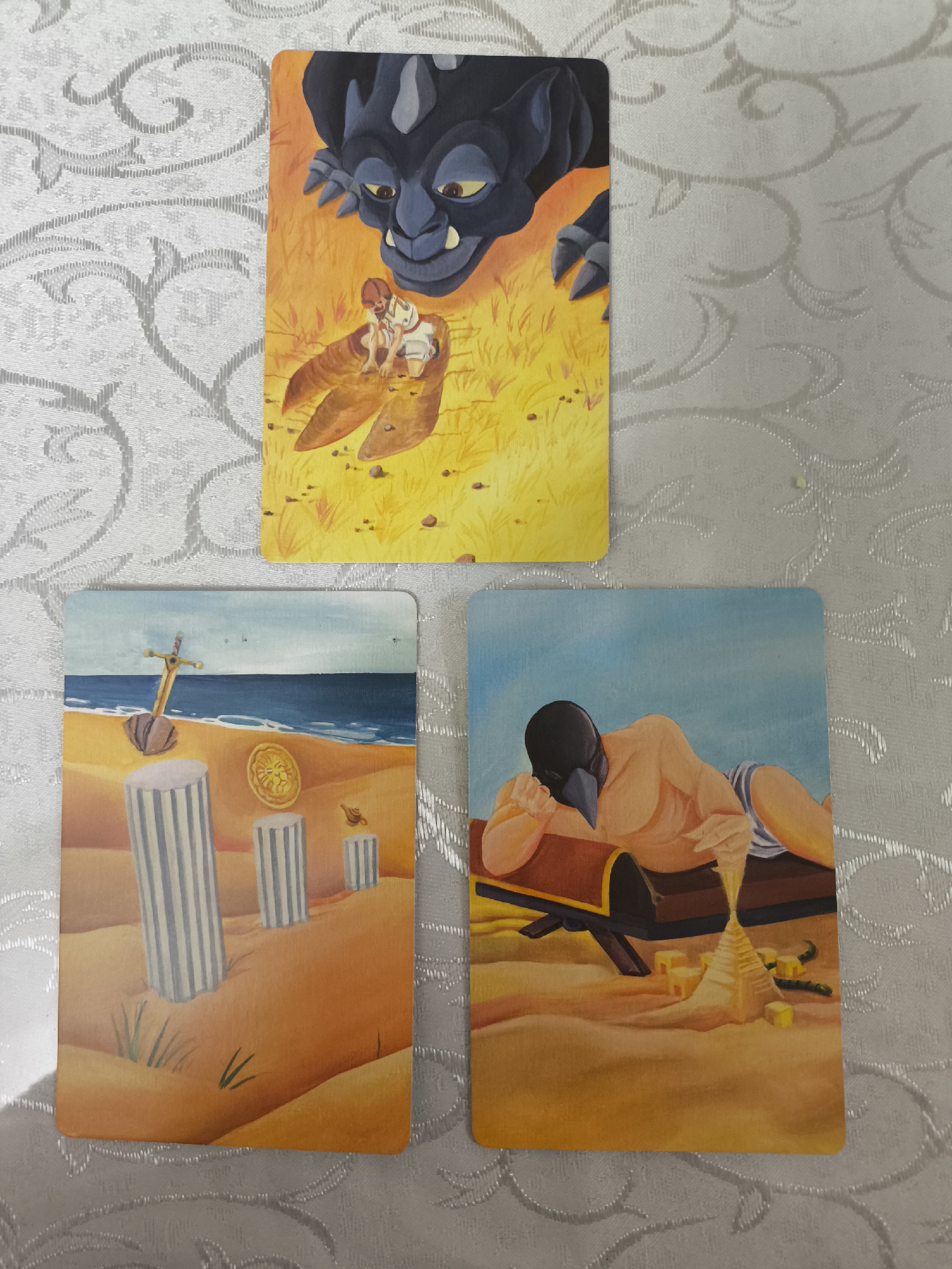 Three tarot cards are laid out on a white patterned tablecloth.
The top card depicts a small, fair-skinned person in white clothes kneeling inside a large claw. A large, dark-colored creature with yellow eyes and large teeth looms over them. The background is yellow and orange, possibly suggesting a desert or grassland environment.
The card on the lower left shows a coastal scene. There are several white pillars on a sandy beach, with a sword stuck in the sand. A gold coin with a lion’s head and a small lamp are also visible. The sea and sky are blue.
The card on the lower right depicts a person with a dark bird-like mask lying on a wooden chest. The person is fair-skinned and wearing striped shorts. In front of the chest are sand blocks forming a pyramid shape, along with some coins. The background is sandy.
There is no indication that this image is located in Izhevsk, Russia.