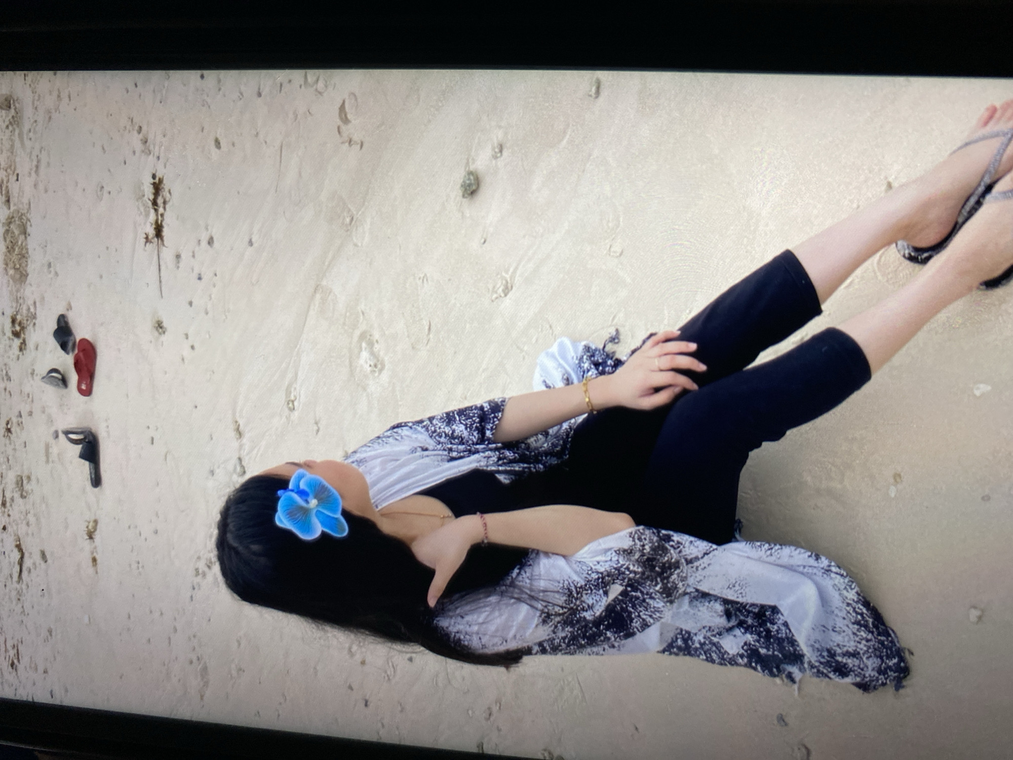 A woman with long, dark hair and a blue flower tucked behind her ear sits on a sandy beach in Ta', Indonesia. She is wearing a black outfit with a patterned kimono-like cover-up and flip-flops. The beach is sparsely populated with a few scattered pieces of debris.